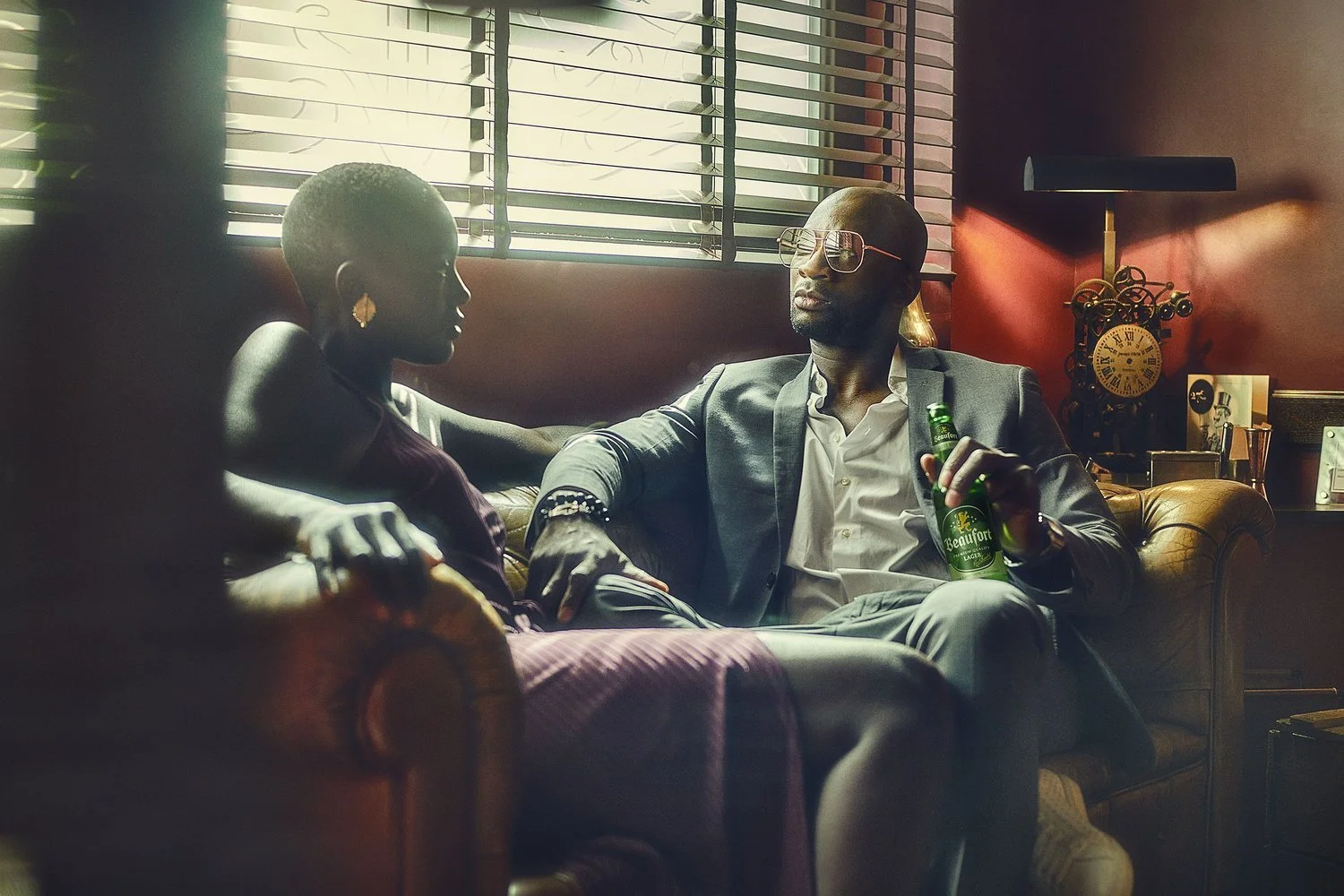 A man and a woman sitting on a leather couch in a dimly lit room with sunlight filtering through blinds, having a conversation. The man is wearing glasses, a light gray suit, and holding a beer, while the woman has a short haircut and large earrings.