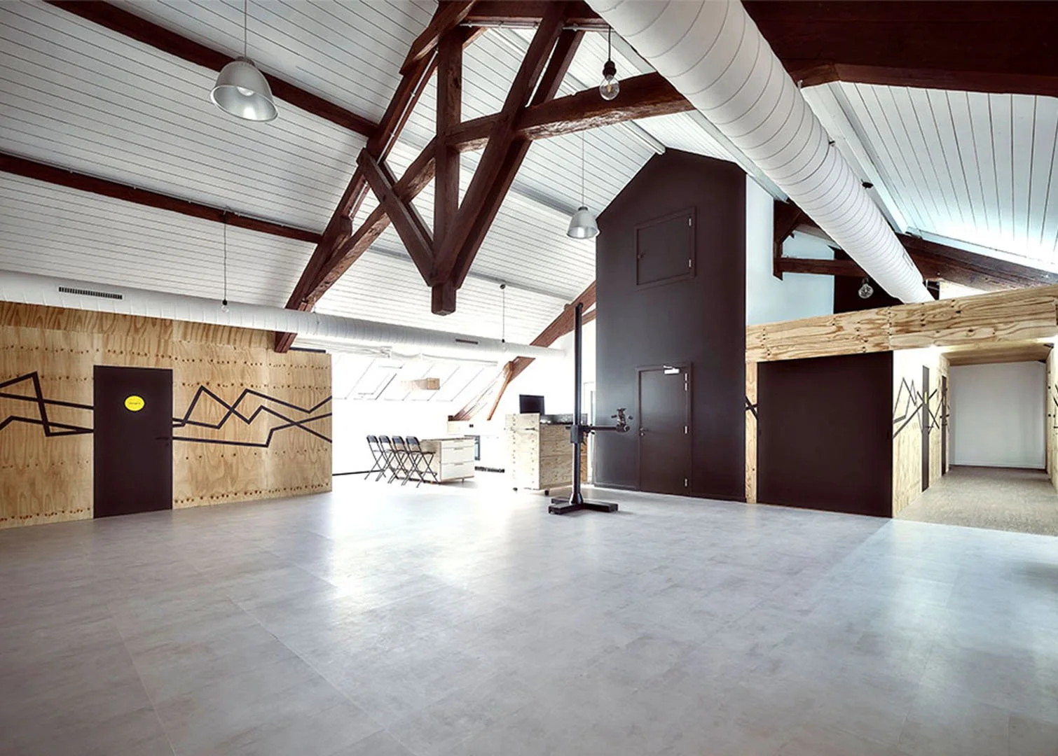 Spacious office or studio space with high vaulted ceiling, wooden beams, and minimal decor. Features a wooden wall with geometric black lines, a black door, and piping on the ceiling. Natural light, a desk with chairs, and some equipment are visible.