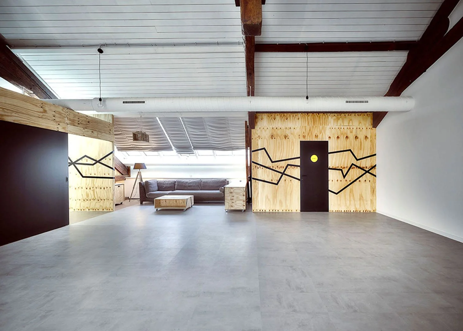 Modern loft with wooden accent wall featuring black geometric design, gray sofa, wooden coffee table, standing lamp, and large windows with skylights