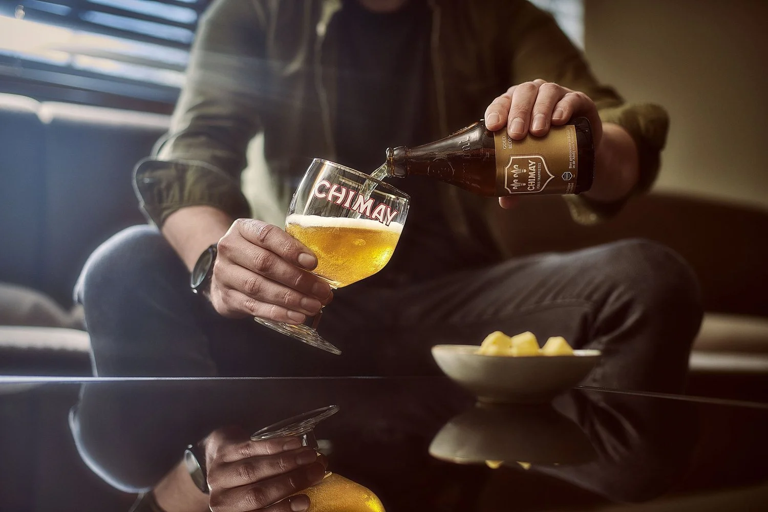 A man sitting on a couch pouring beer from a Chimay bottle into a Chimay branded glass