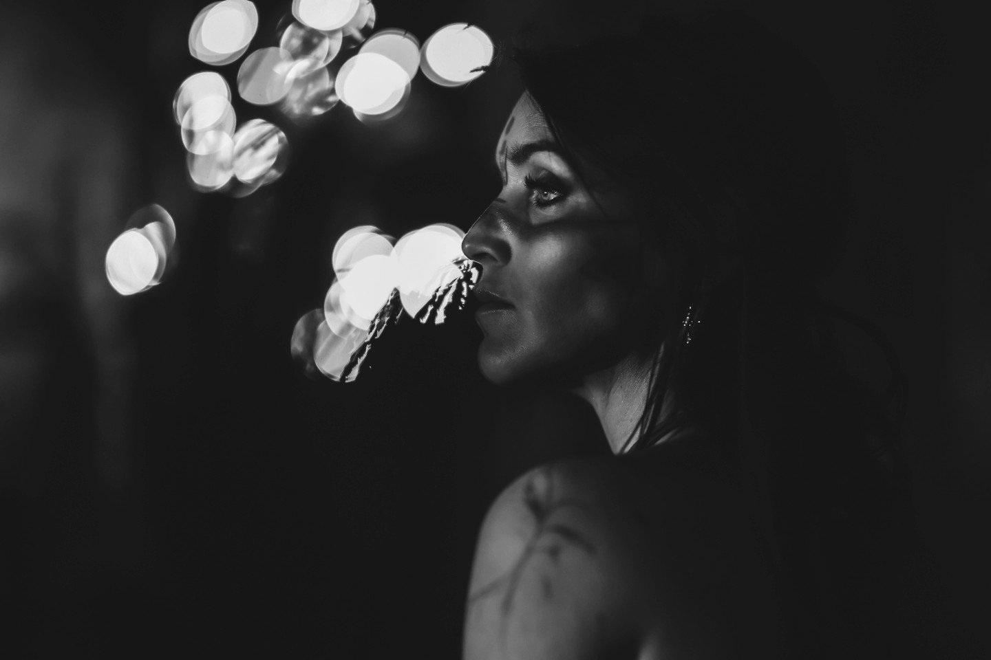 A black and white close-up photo of a woman with earrings, looking up and to the right, with bokeh lights in the background.