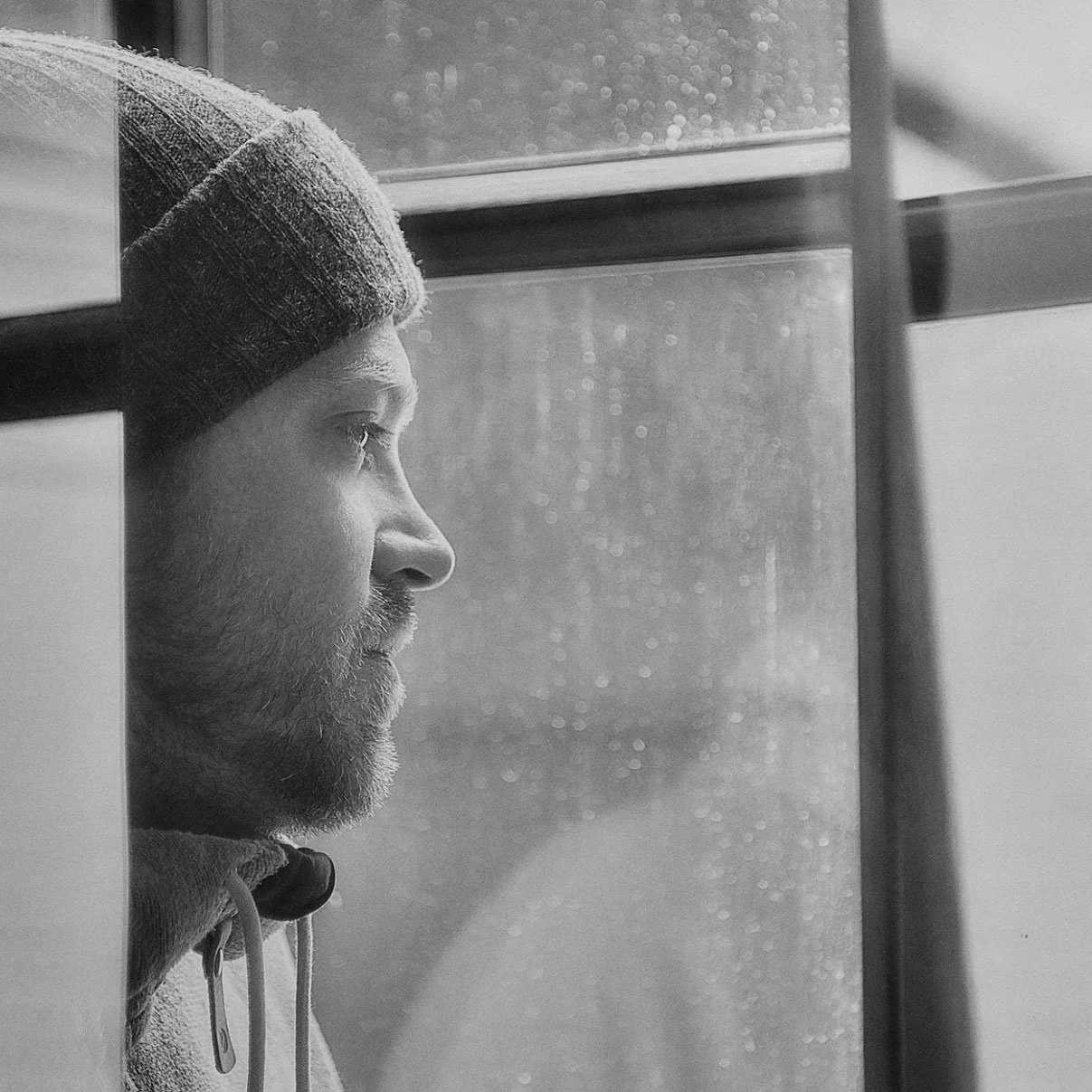 A man with a beard wearing a beanie, looking out of a window in black and white.