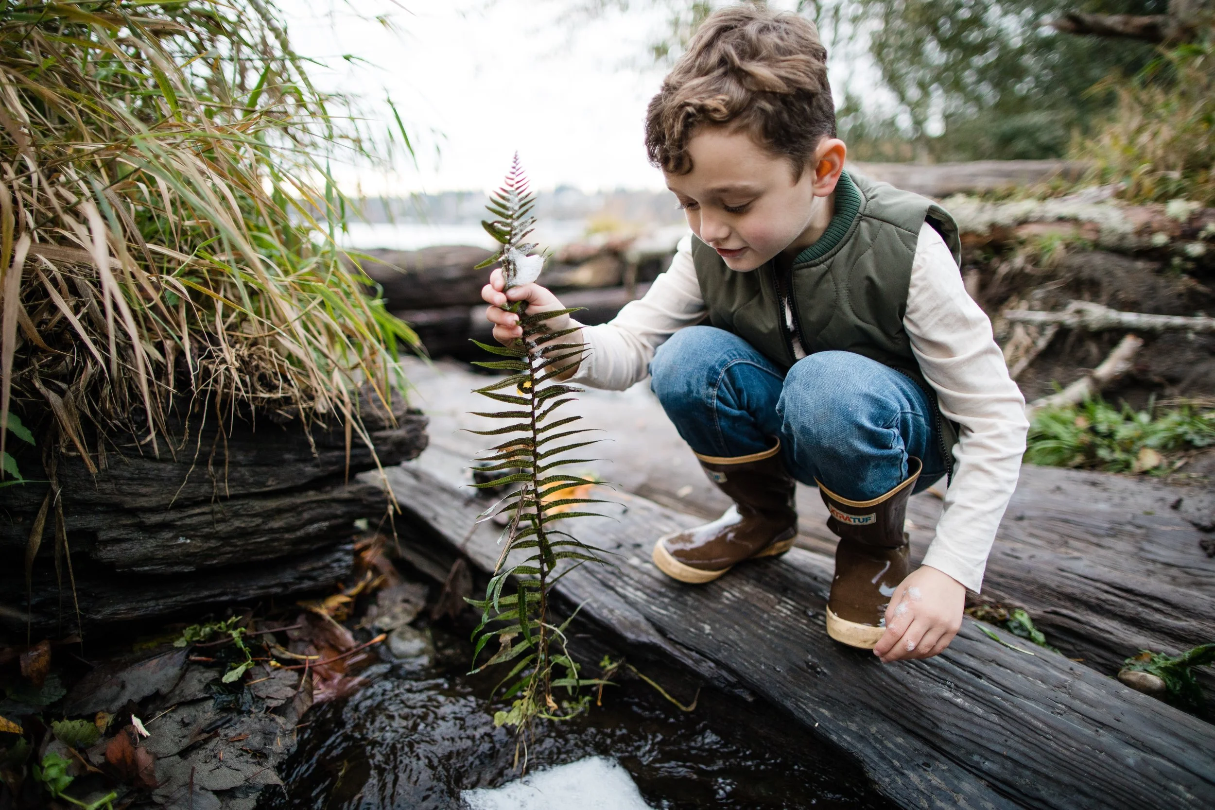 A boy plays with a branch while standing on a log. It has just rained and the ground is wet.