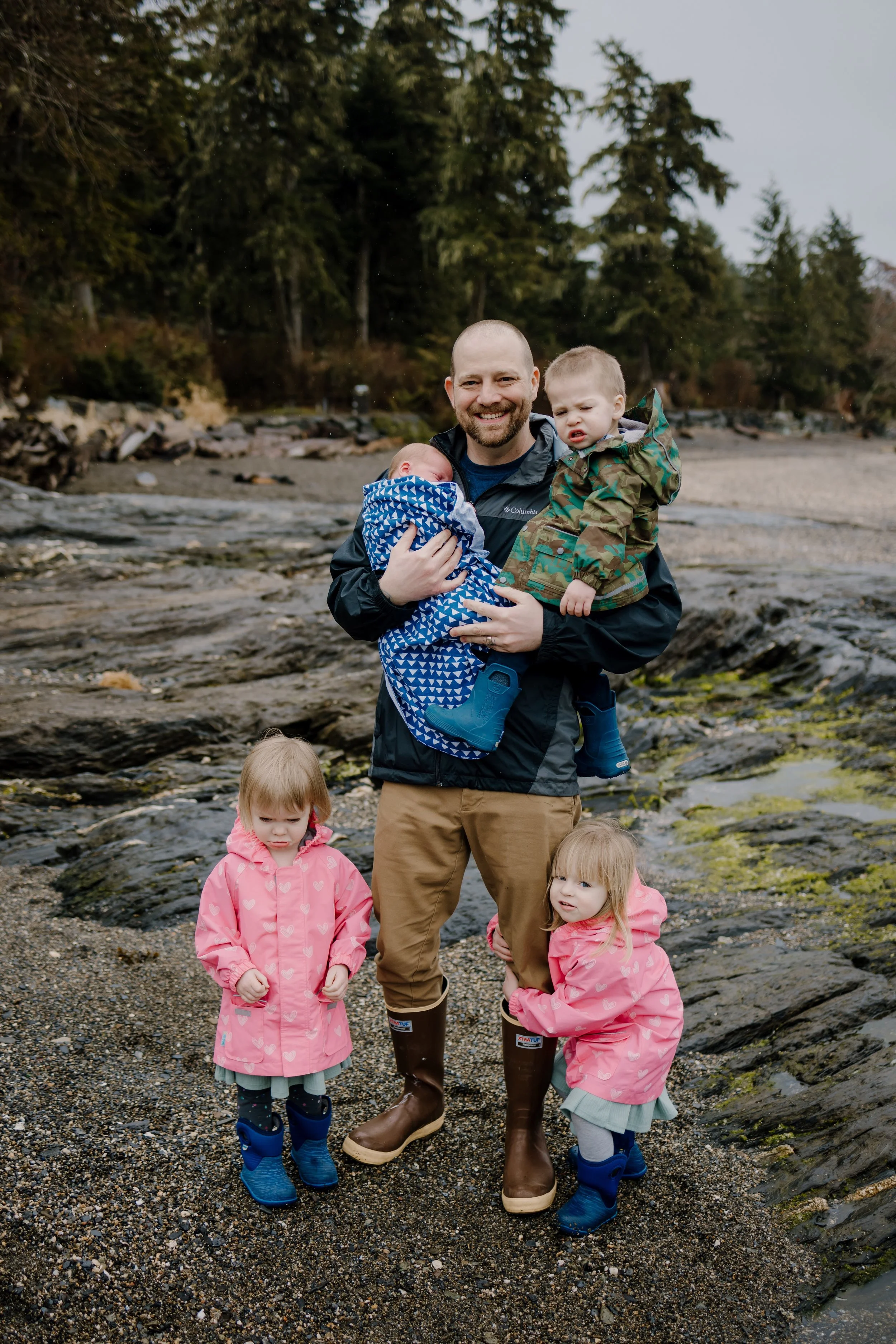 A father holds his toddler and infant sons while his daughters are standing near his legs. They are on a beach during low tide and they are wearing rain coats and boots.