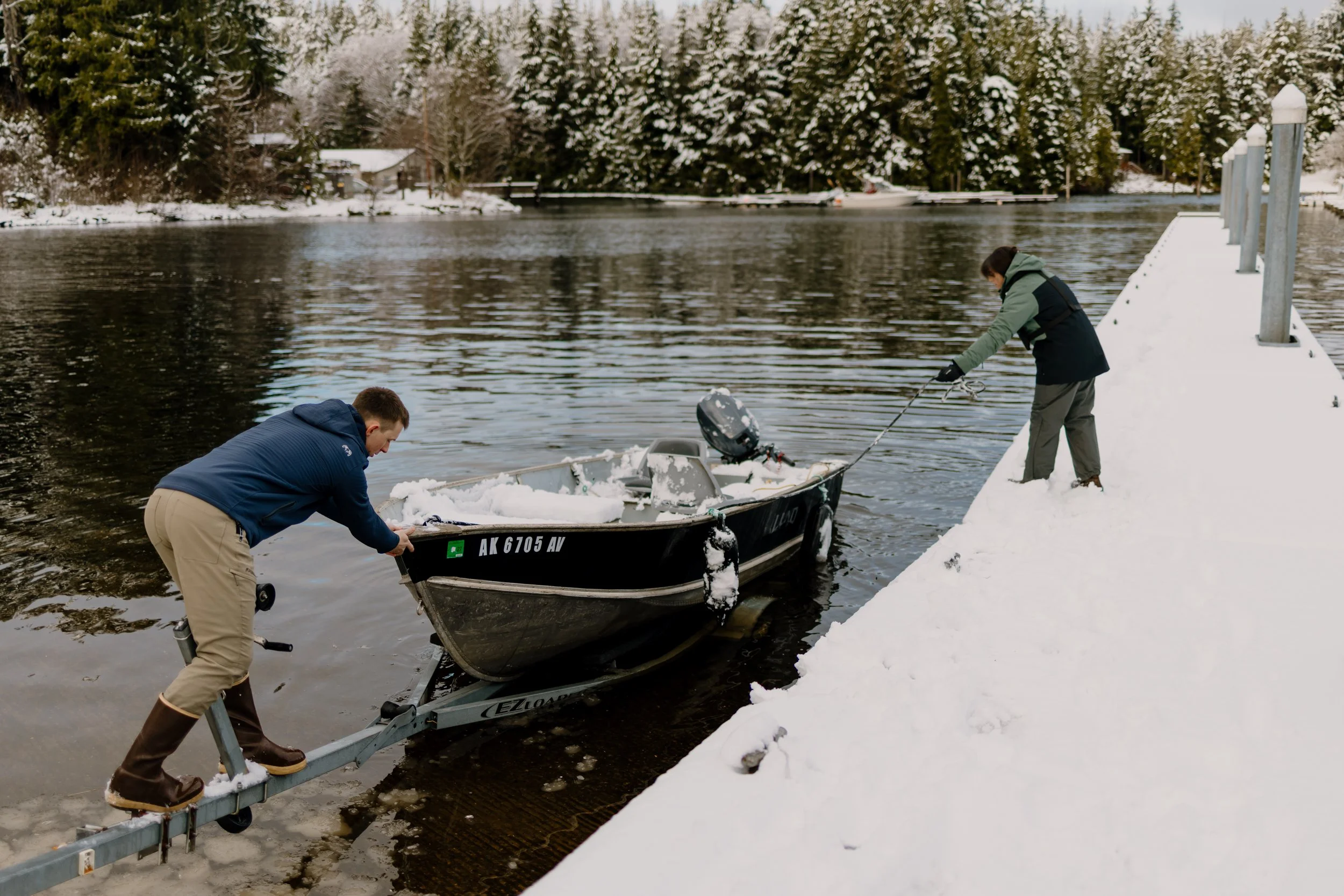 A dad and his son are pushing a small boat into the water. The dad is standing on the trailer and the son is standing on a snowy dock.
