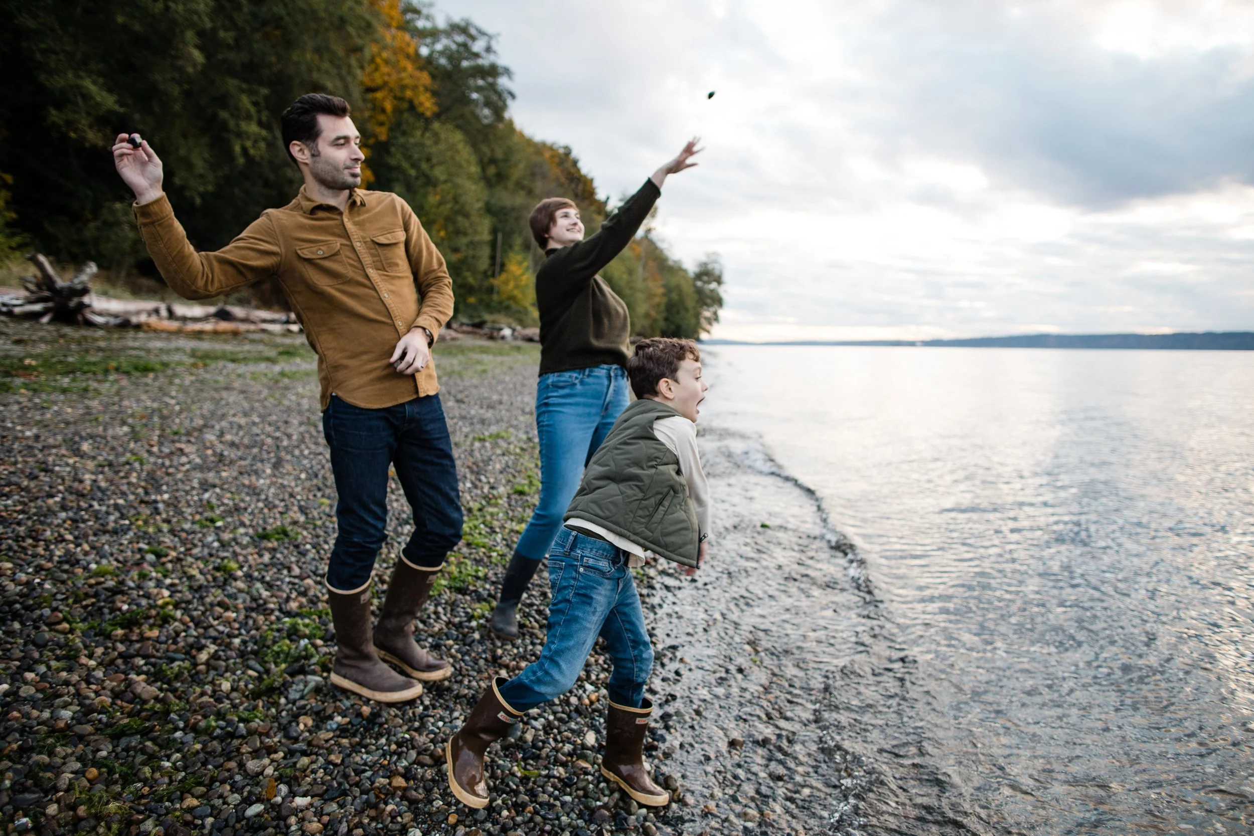 A dad, mom, and son are throwing rocks into the water on a rocky beach. They are wearing xtratuf boots and fall clothing. The sky is cloudy.
