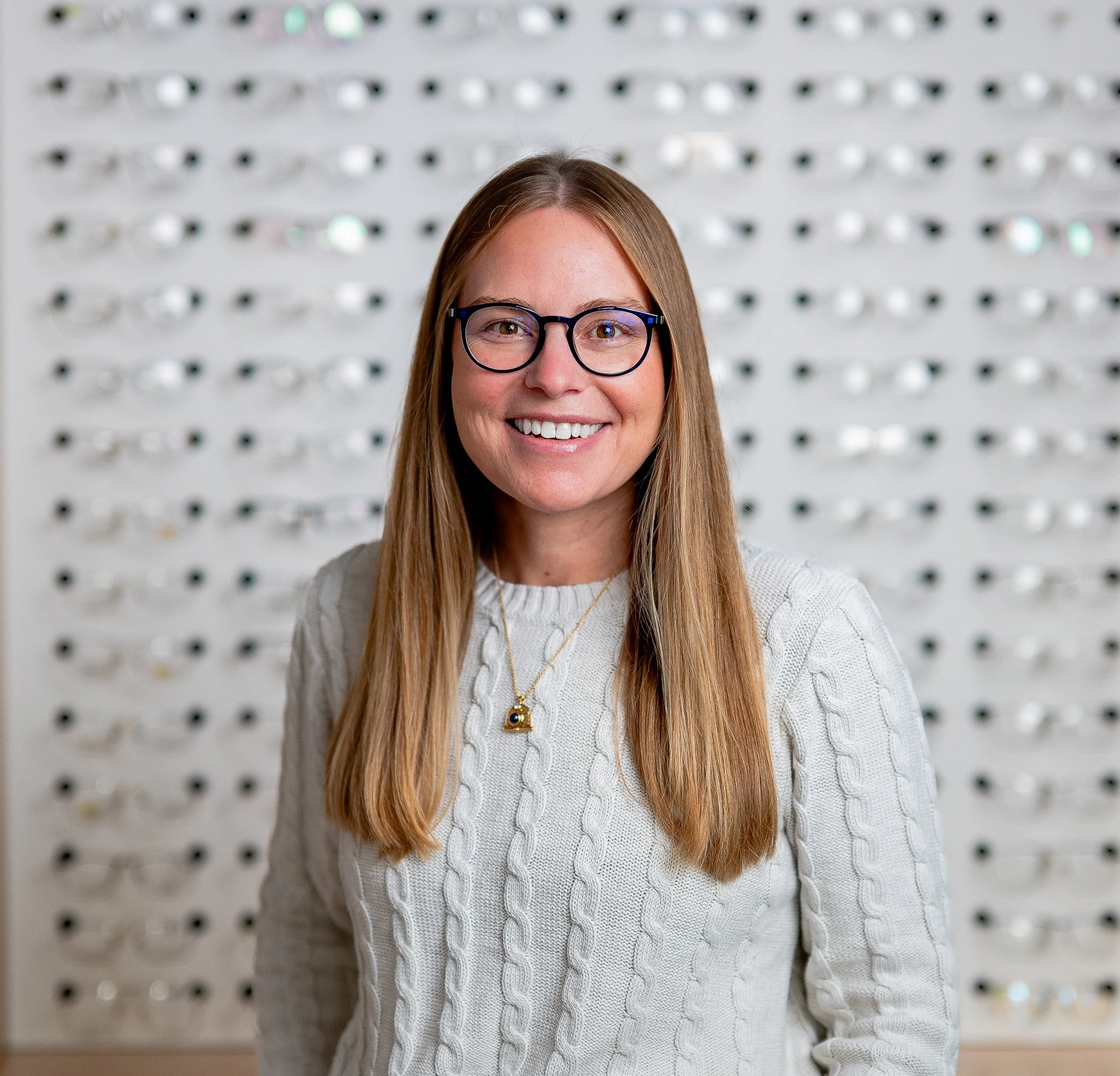 A woman with long, straight, light brown hair, wearing glasses and a light beige sweater, smiling in front of a display of eyeglasses.