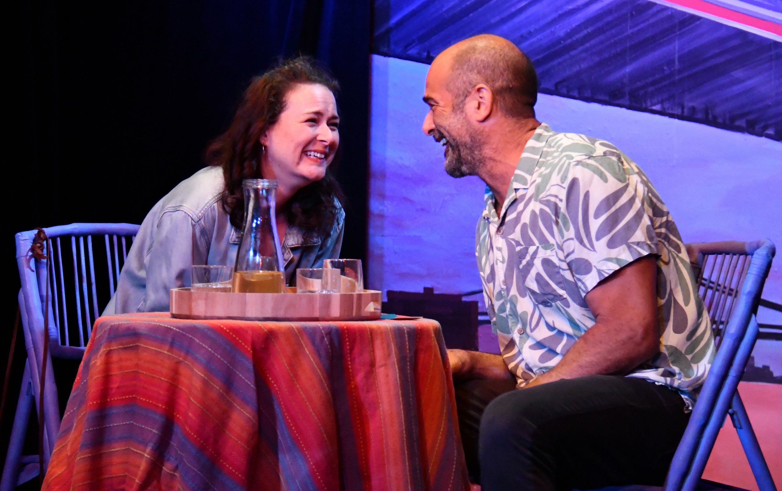 A woman and a man share a joyful moment at a table indoors with purple lighting, with drinks and a bottle on the table, and a brick wall in the background.