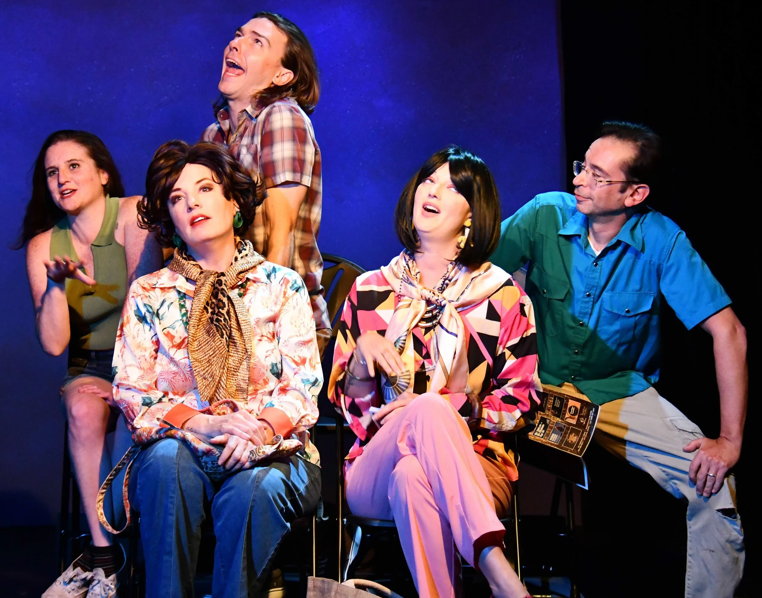 Group of five actors performing on stage, dressed in colorful costumes, with a plain blue background.