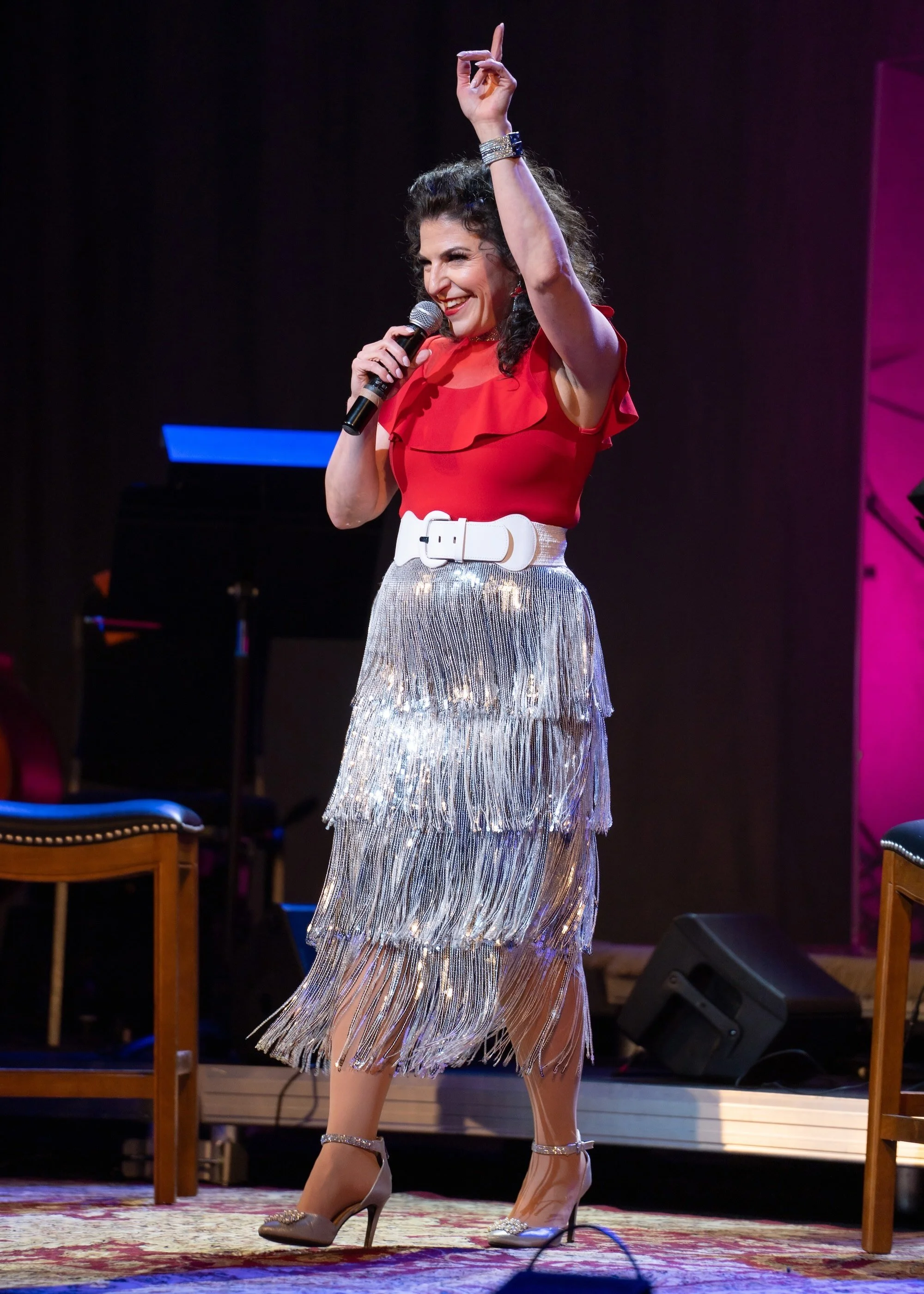 woman wearing a red top and silver sparkly skirt holding mic pointing to the sky