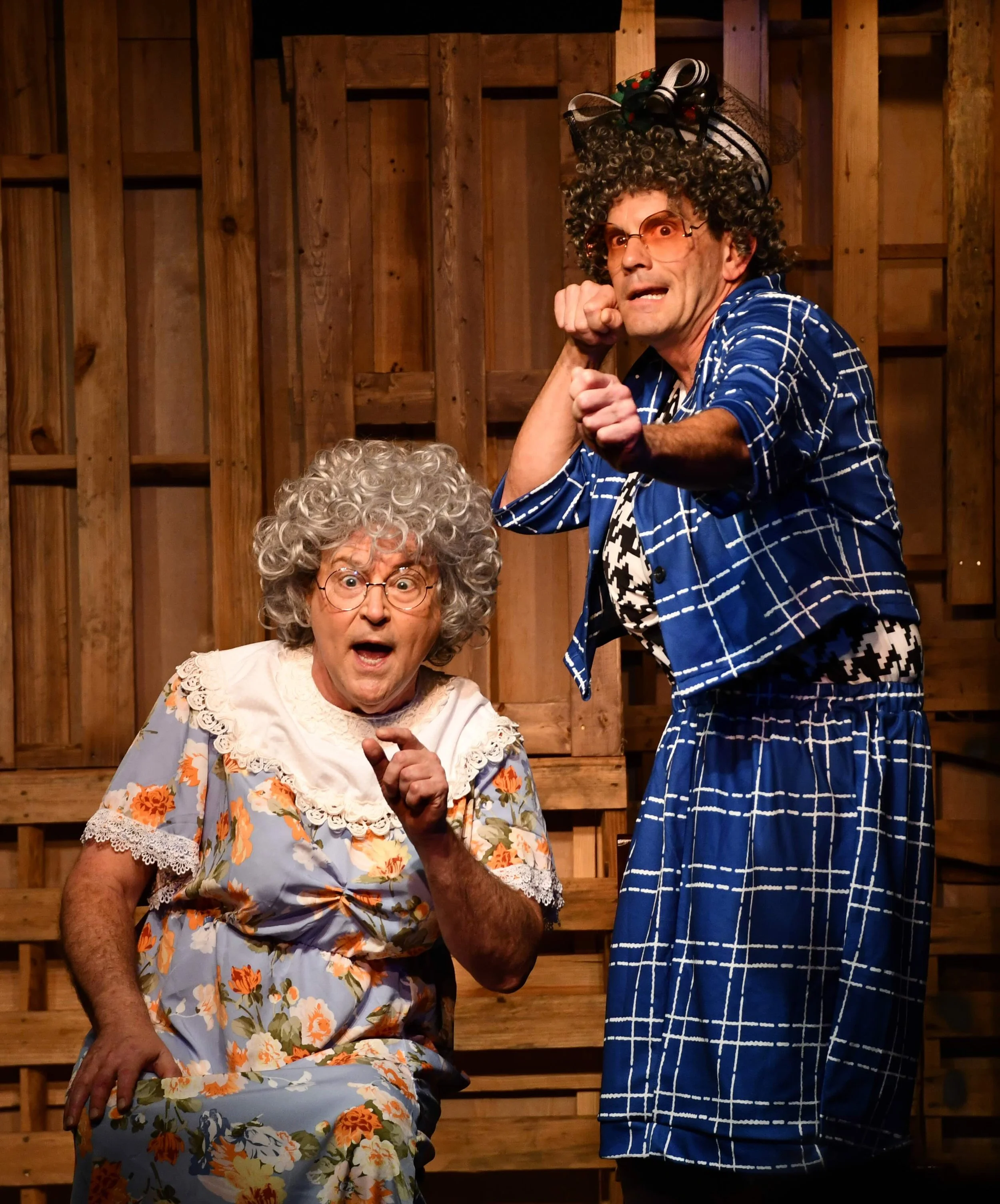 Two older women and a man are in a wooden room. The woman in the foreground is elderly, wearing glasses and a floral dress, with a surprised expression. The man behind her is wearing a hat decorated with flowers, glasses, and a blue checkered outfit, making a dramatic gesture. The background consists of wooden panels.