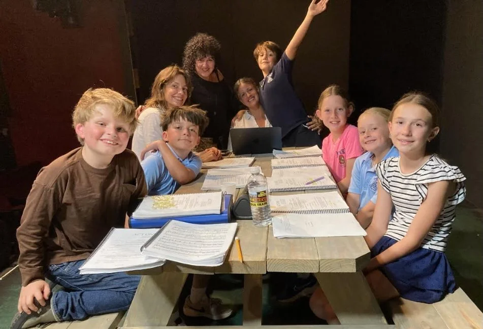group of children smiling and sitting at a long picnic table with books and scripts in front of them