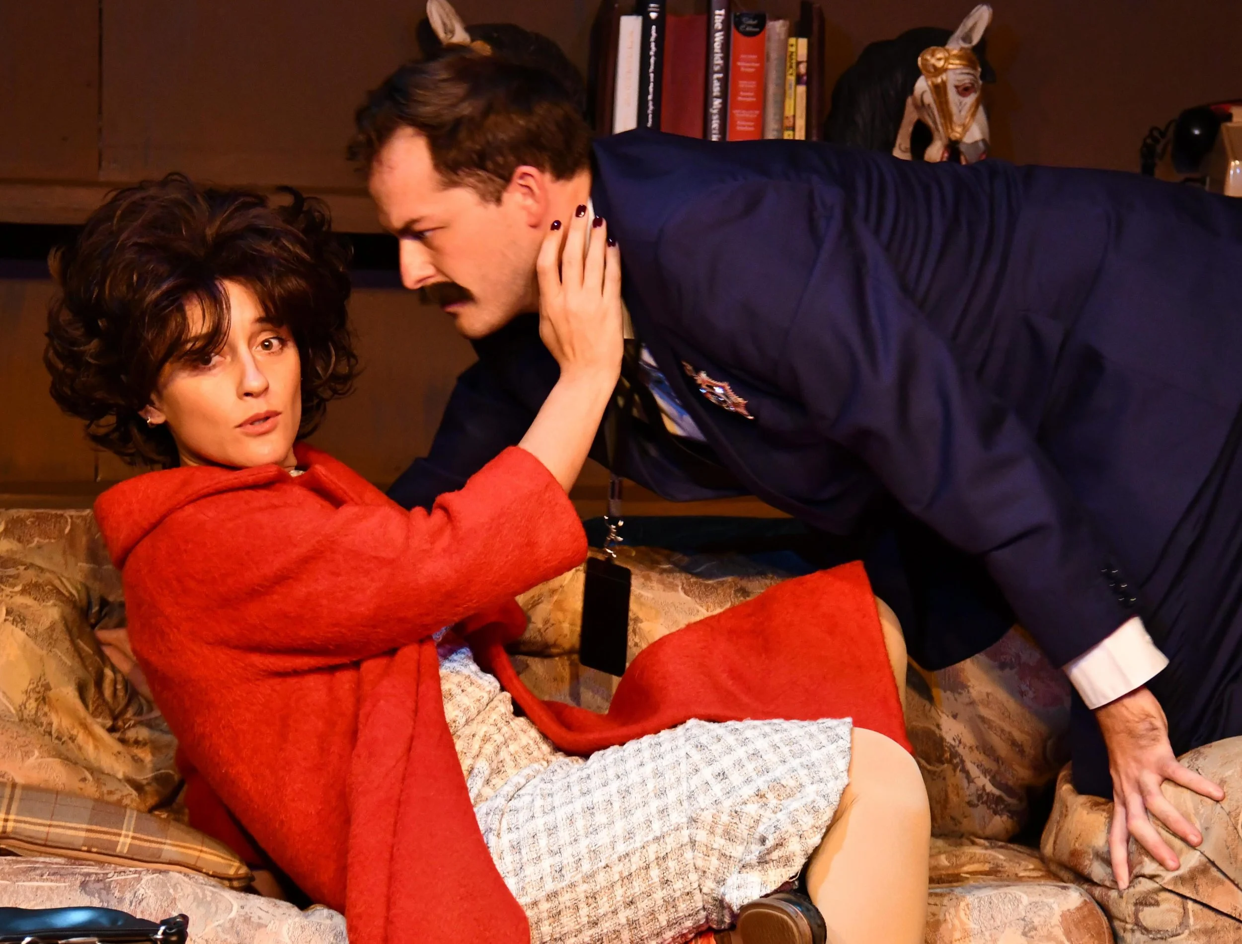 A woman in a red sweater sitting on a sofa, looking surprised, while a man in a dark jacket leans over her with his face close to hers, touching her cheek with his hand. There are books and decorative items on a shelf behind them.