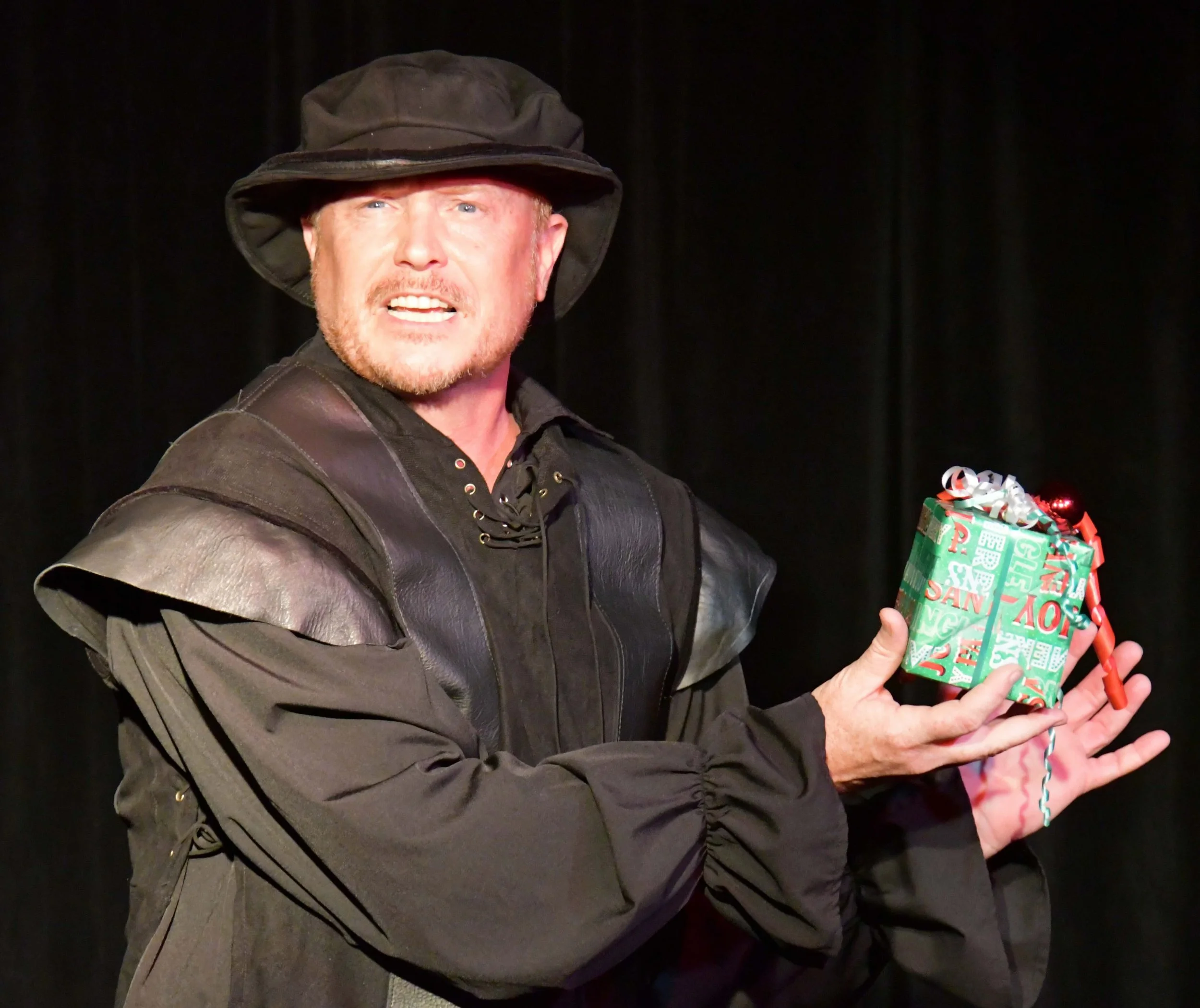 A man with a reddish beard and blue eyes wearing a black hat and black leather jacket, holding a wrapped Christmas gift with colorful paper and ribbons.