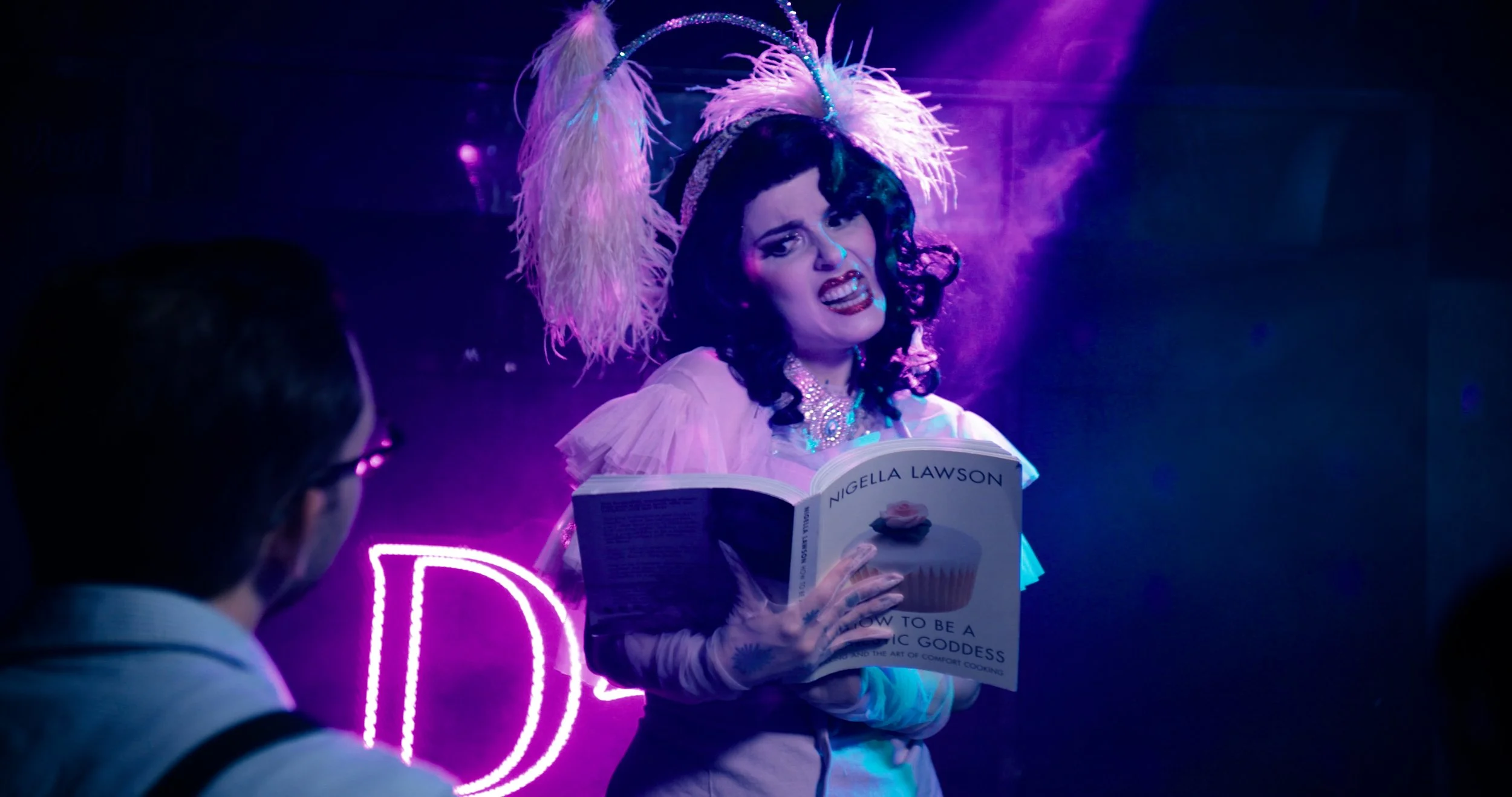 Still from a documentary series "KulturDocs," featuring an actor in a costume with a book entitled Nigella Lawson