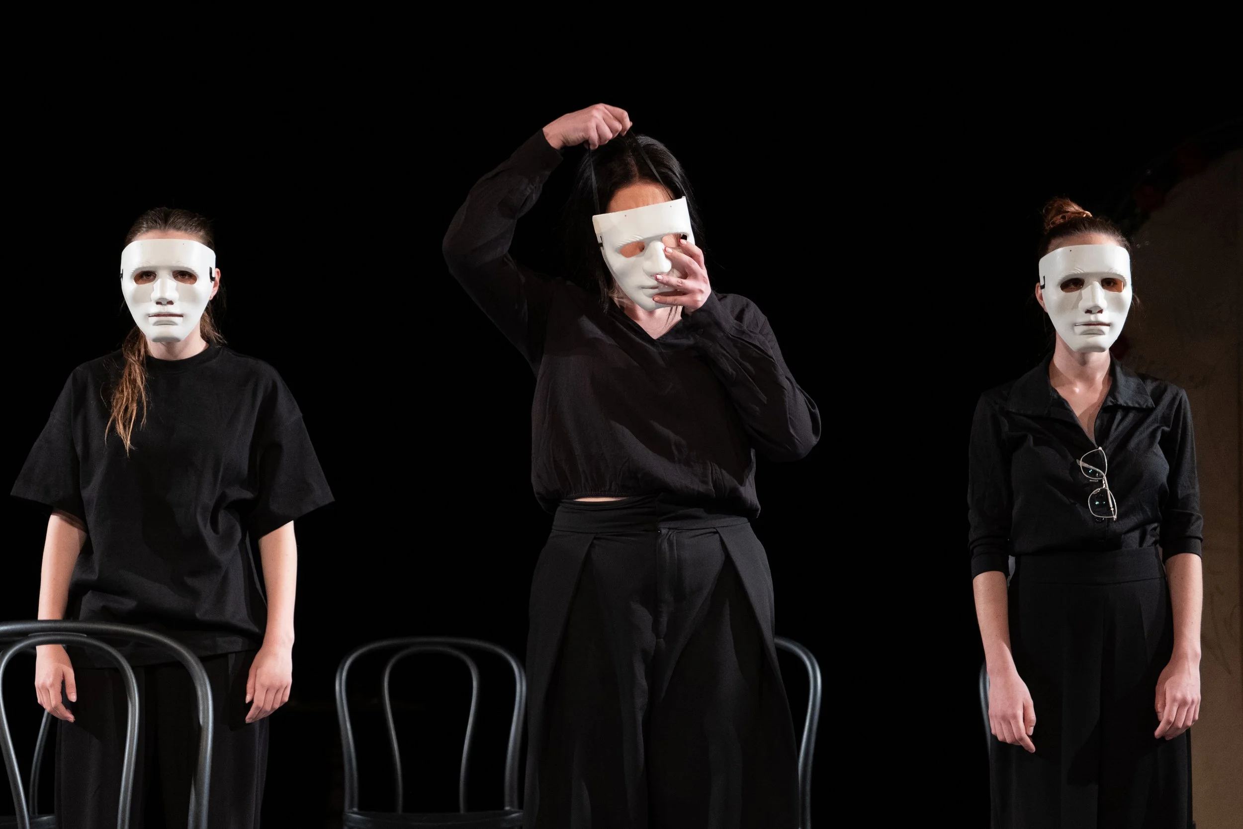 Three actors with white masks on standing around black chairs.