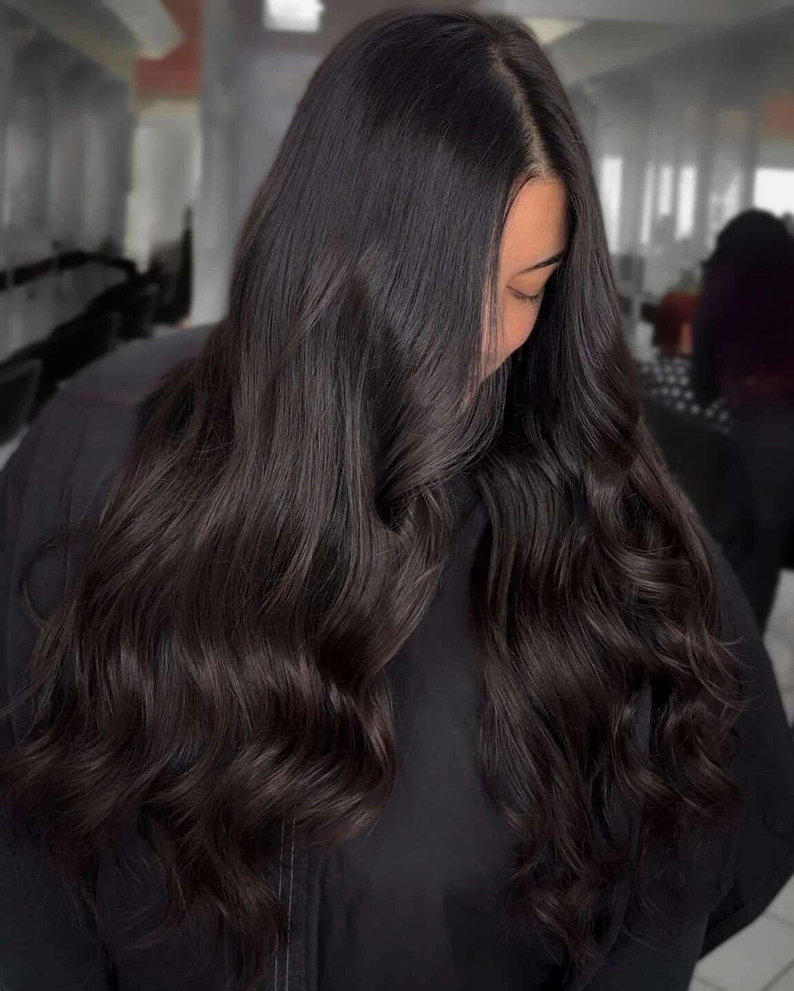 Hair extensions result photo Bergen County NJ salonA woman with long, dark wavy hair, wearing a black top, looking downward in an indoor setting with large windows.