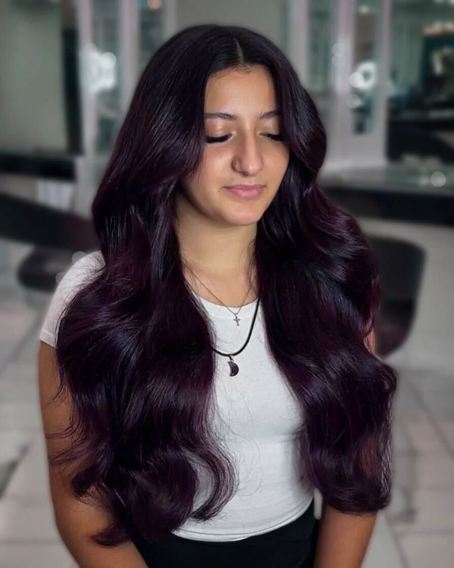 Color-matched hair extensions at Bellibone Salon Maywood NJA young woman with long, dark, wavy hair with purple highlights, wearing a white t-shirt, small cross and moon necklaces, standing in a salon or modern indoor space with her eyes closed.