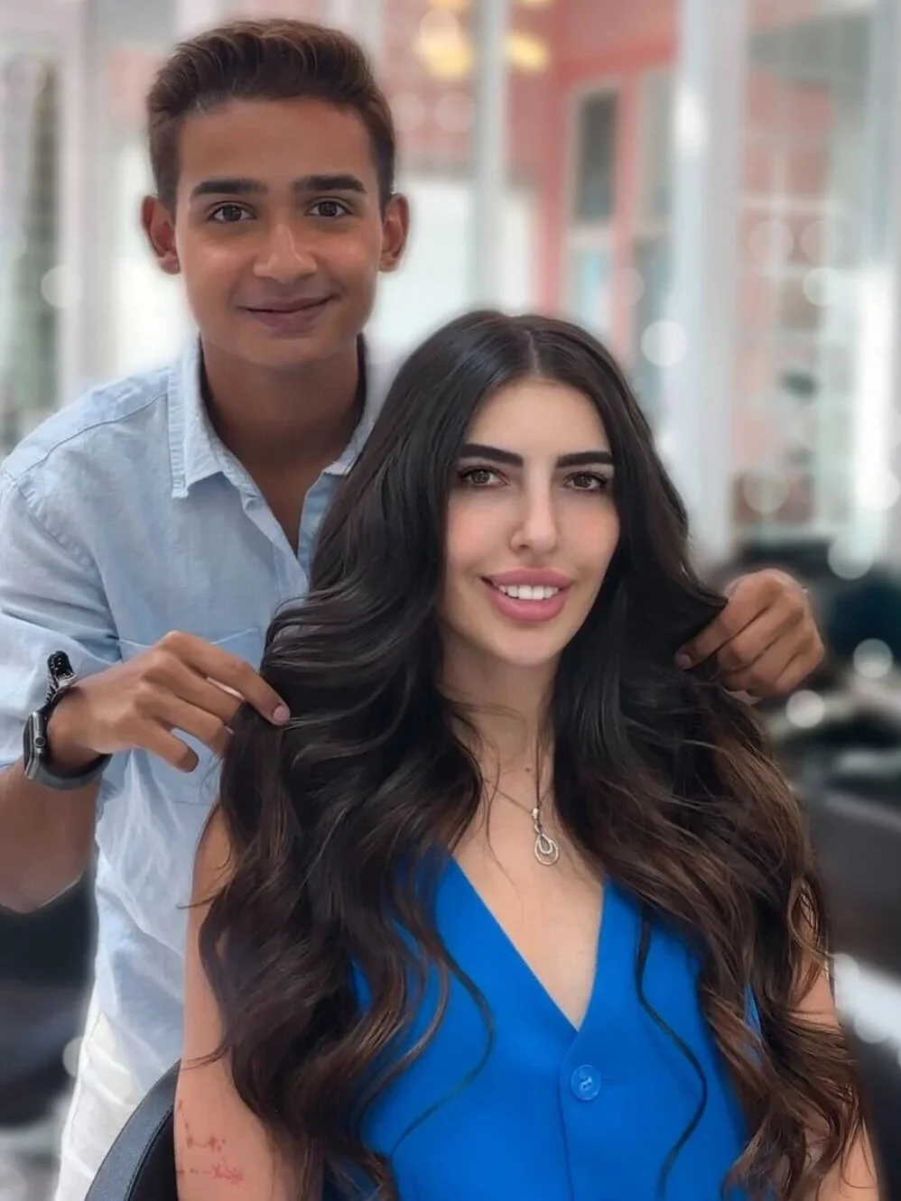 Fashion hair color and extensions at Bellibone Salon Bergen County NJA hairstylist with short brown hair, smiling, standing behind a woman with long, wavy dark hair, wearing a blue top, and holding a section of her hair.