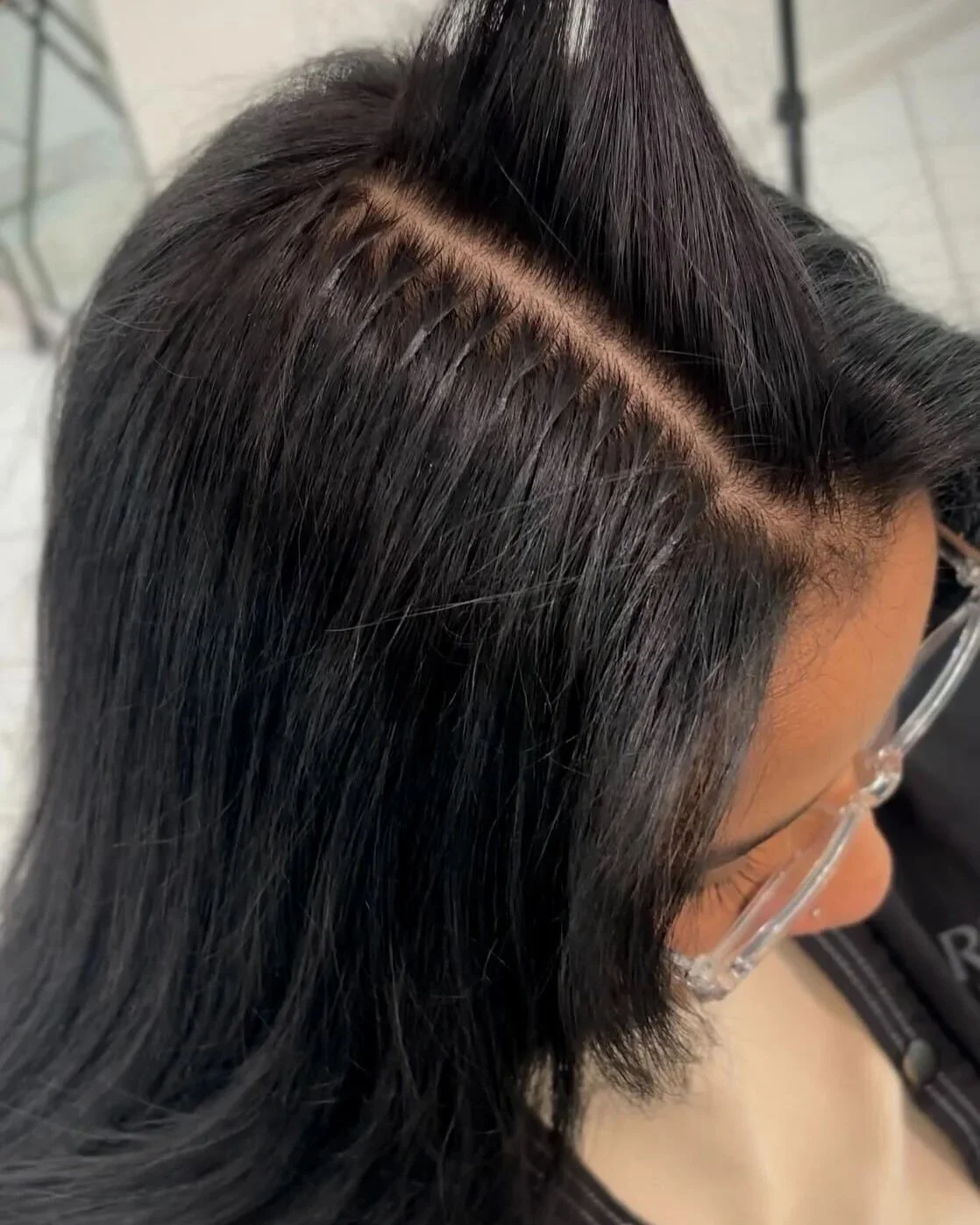 Hand-tied weft hair extensions for volume and length in Maywood NJClose-up of a woman's hairstyle showing a straight part with sleek, black, shoulder-length hair and visible scalp.