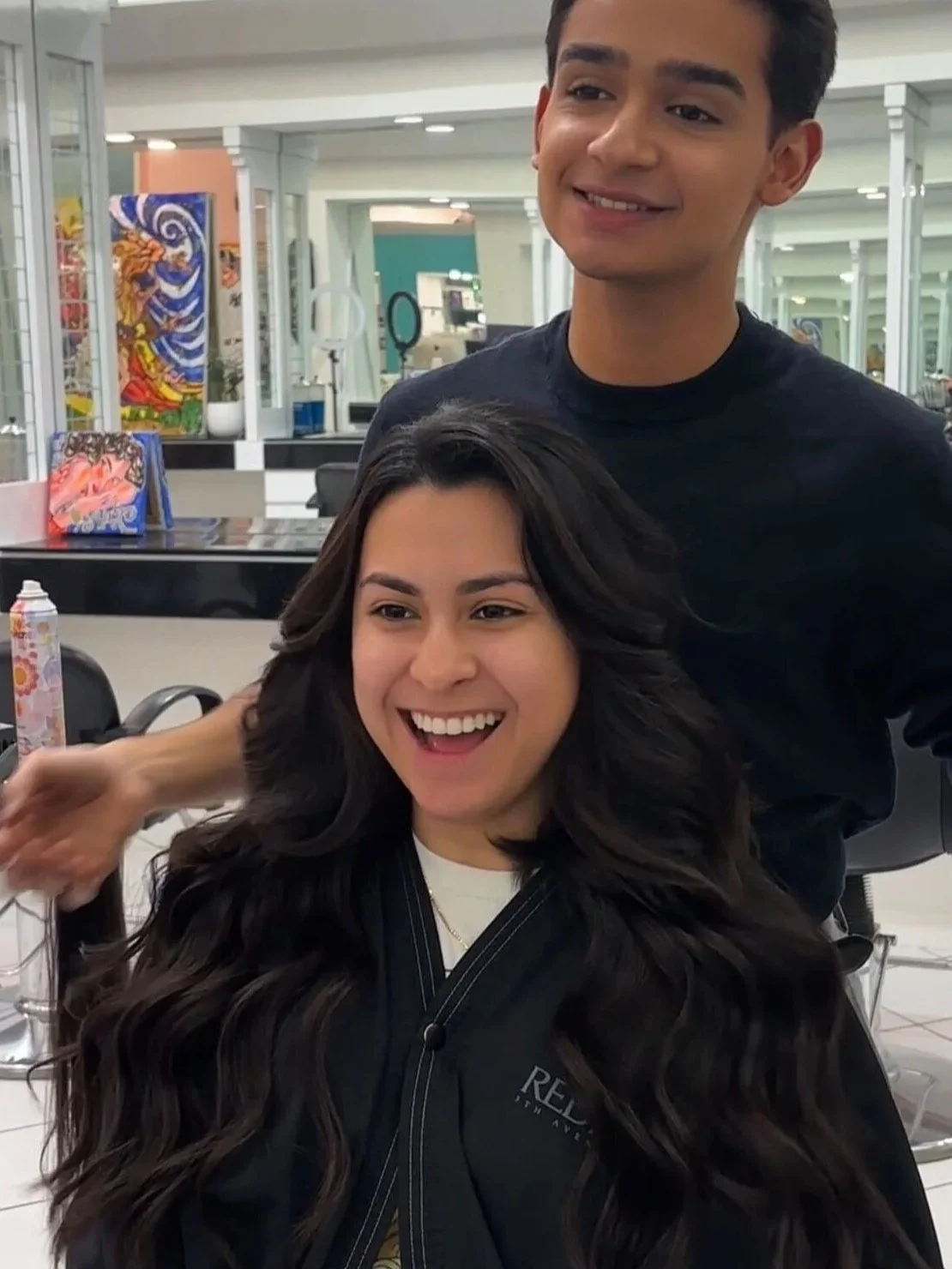 Hair extensions length and volume result at Bellibone Salon Bergen CountyA woman with long, wavy dark hair sitting in a salon chair, smiling, with a stylist standing behind her, in a bright salon with colorful artwork on the walls.