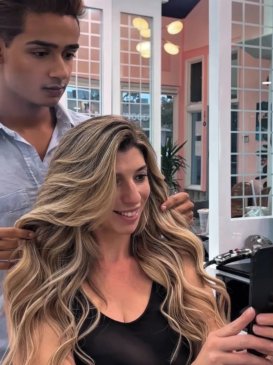 Hair extensions styling result at Bellibone Salon Maywood NJA woman with long, wavy blonde hair in a black top takes a selfie while a stylist in the background prepares her hair in a salon with pink walls and large windows.
