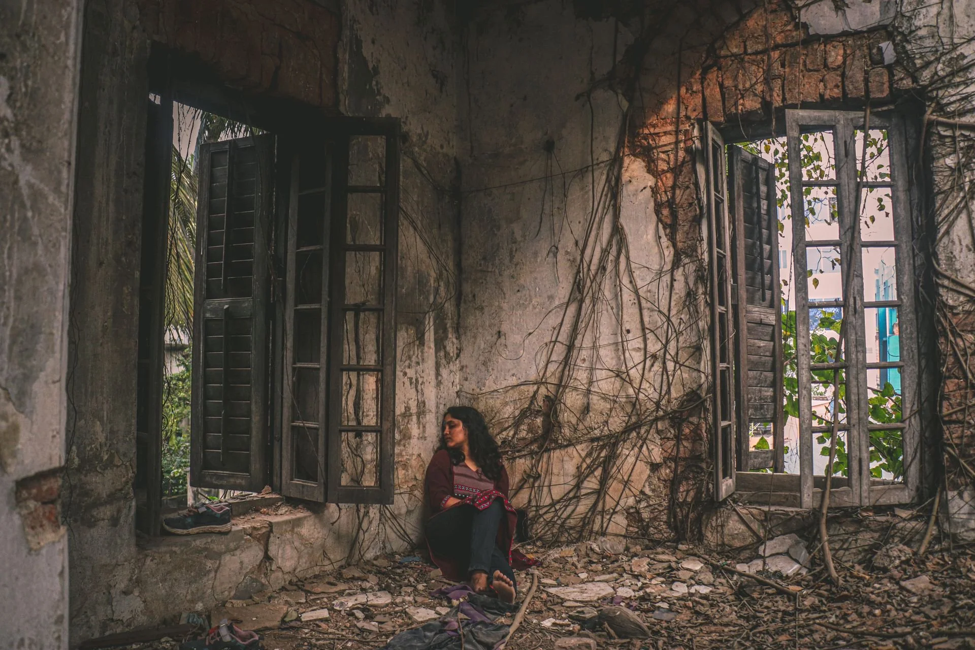 emotional and sad woman sitting in between two large open windows in abandoned room with vines growing up the dilapidated walls