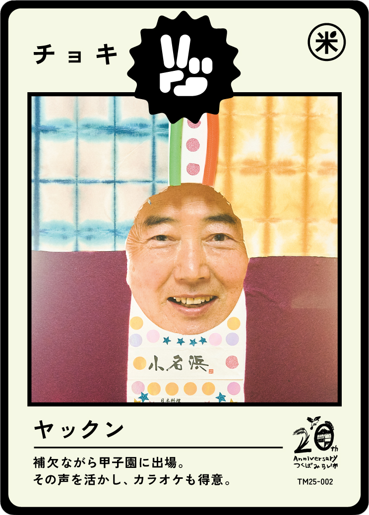 A humorous photo of a man’s face inserted into a paper puppet costume with colorful patterns, set against a background of multicolored curtains, resembling a card from a Japanese game or card set.