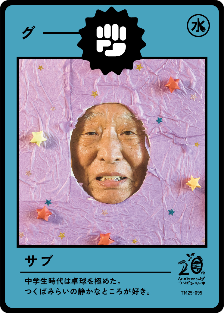 A person with a visibly toothless smile looking through a cut-out hole in a pink paper decorated with small stars and animals, resembling a trading card or collectible card with Japanese text and graphics.
