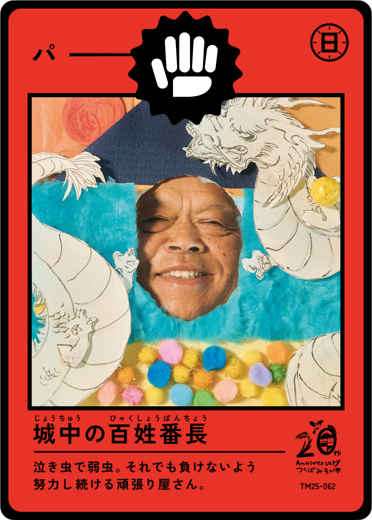 A colorful illustration of a man smiling with his face appearing through a cutout in a decorative display featuring a dragon, colorful pom-poms, and Japanese text.
