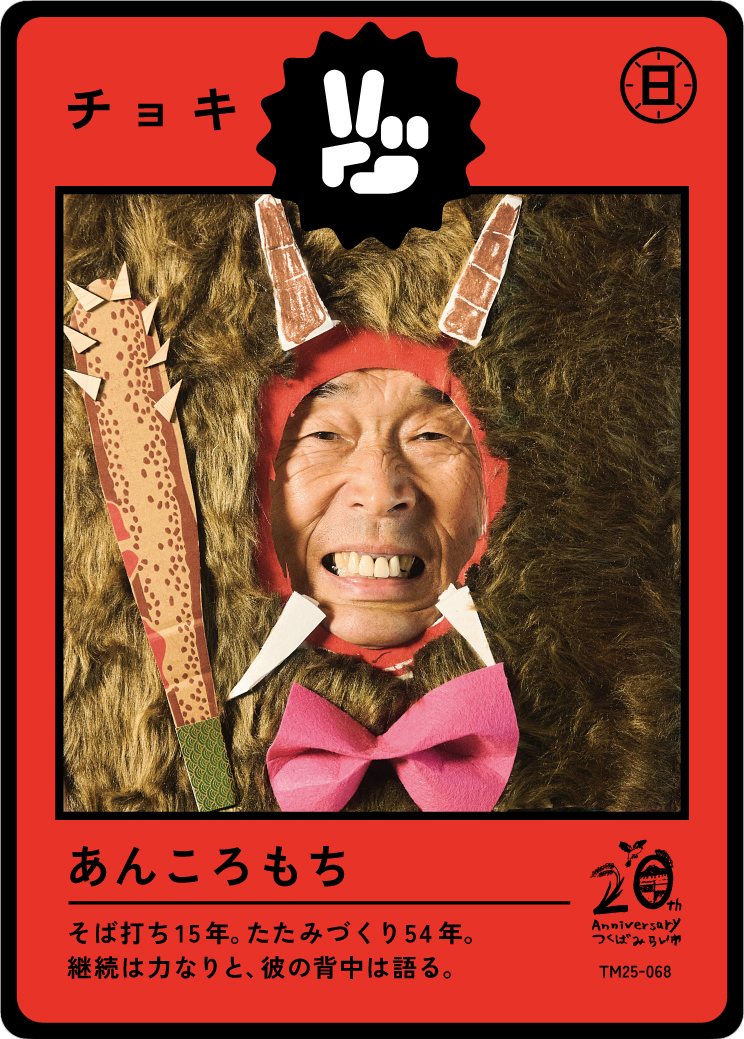 A person with a human face is inside a costume that looks like a creature with horns, large furry body, and a pink bow. The costume has a circular opening around the face. The background is red with Japanese text and symbols, including a black and wh