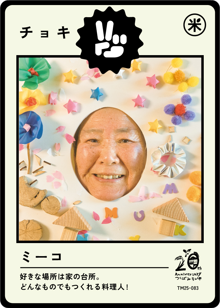 A person looks through a colorful, decorated paper frame with a face cutout. The frame is decorated with small, colorful balls, paper stars, wooden shapes, and alphabet letters. The frame has Japanese text and a 20th anniversary logo.
