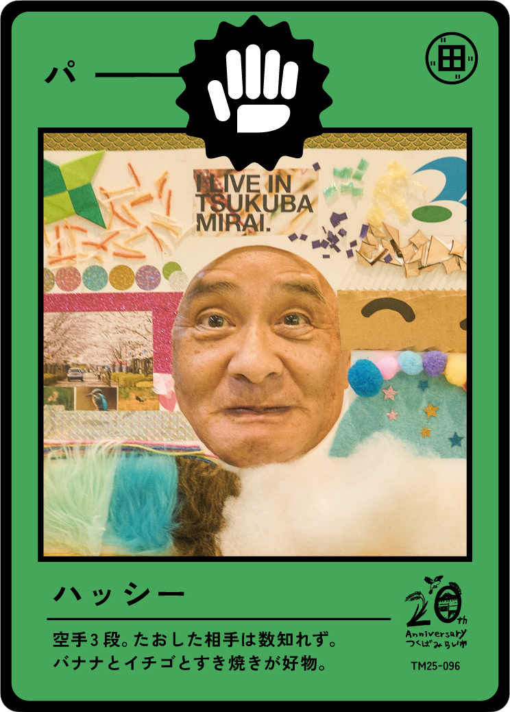 A person taking a selfie in front of a colorful display with paper crafts, photographs, and decorations at Tsukuba Mirai. The display includes the words "Live in Tsukuba Mirai" and various artistic elements.