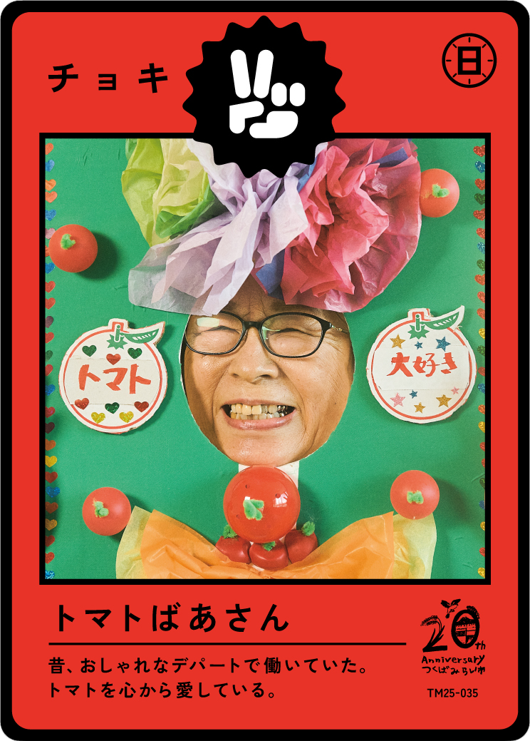 A person wearing glasses has their face visible through a cutout in a colorful, tomato-themed craft display with a large paper hat and decorations, including tomatoes and Japanese text, celebrating an anniversary.