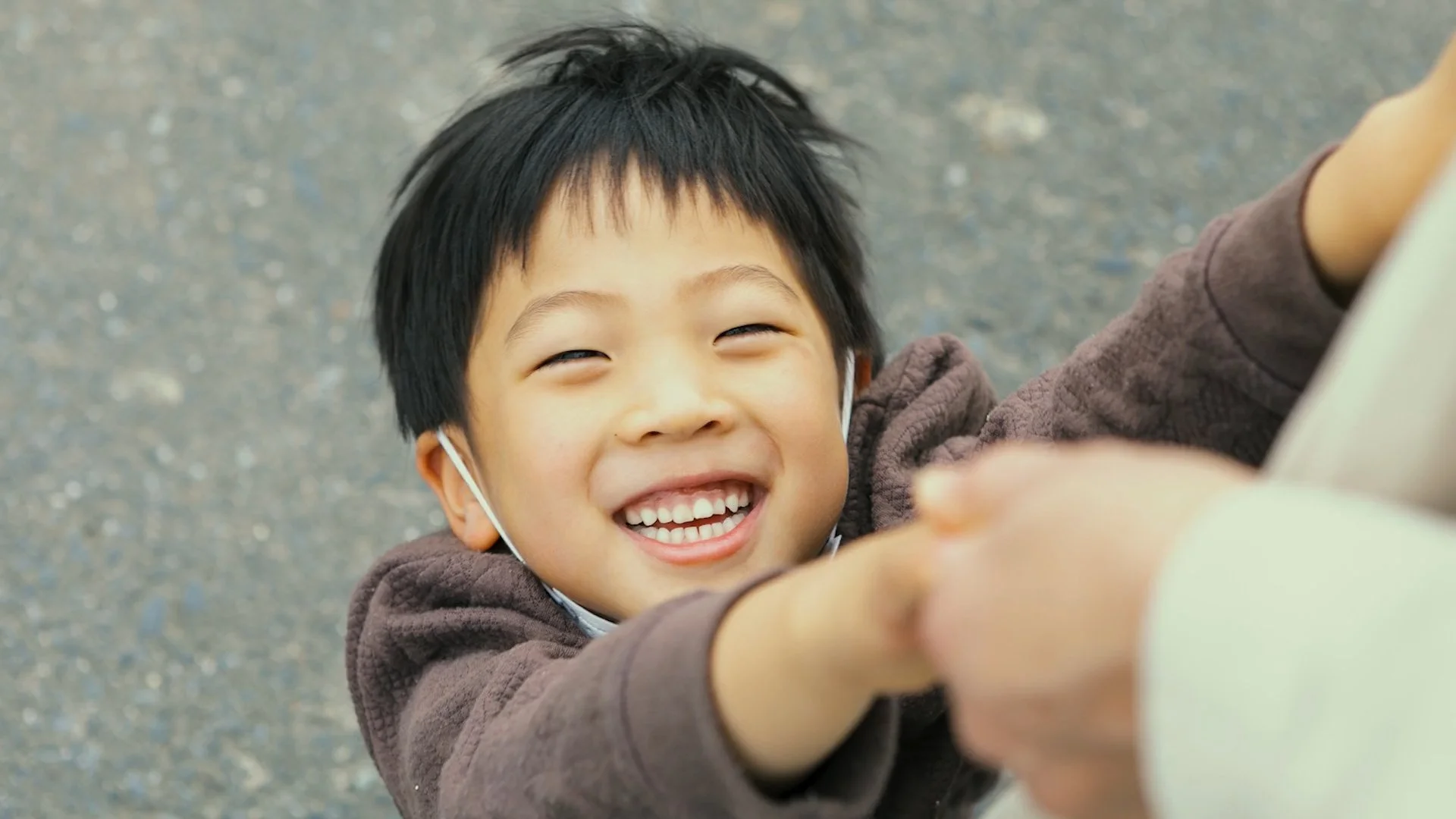 A young boy with black hair smiling happily, reaching out to hold someone's hand outside on a paved surface.