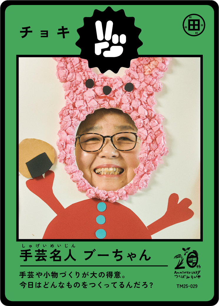 A person with glasses smiling through a photo frame decorated with pink tissue paper resembling a cartoon character, holding a piece of craft material.