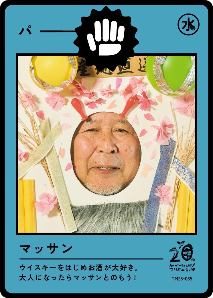 Person with a face cutout in a decorative frame resembling a festive or humorous card, decorated with pink paper flowers, balloons, and streamers, celebrating an anniversary, with Japanese text and colorful design elements.