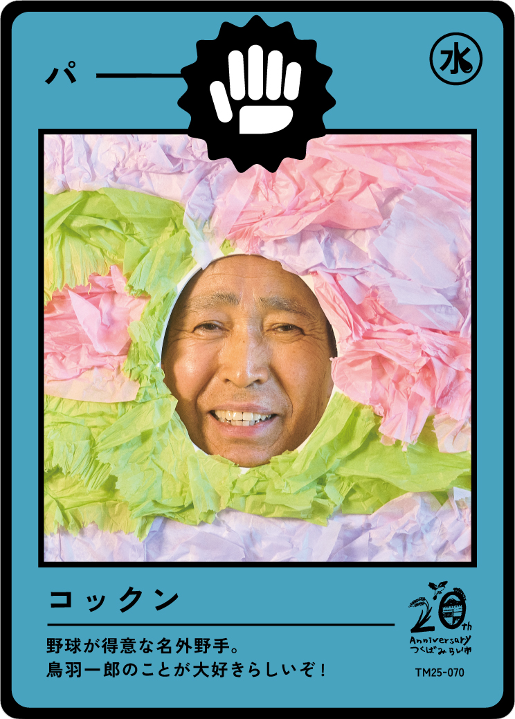 A person’s face peering through a hole surrounded by colorful tissue paper, resembling a comic book character or superhero with a safety icon sticker at the top and Japanese text at the bottom.