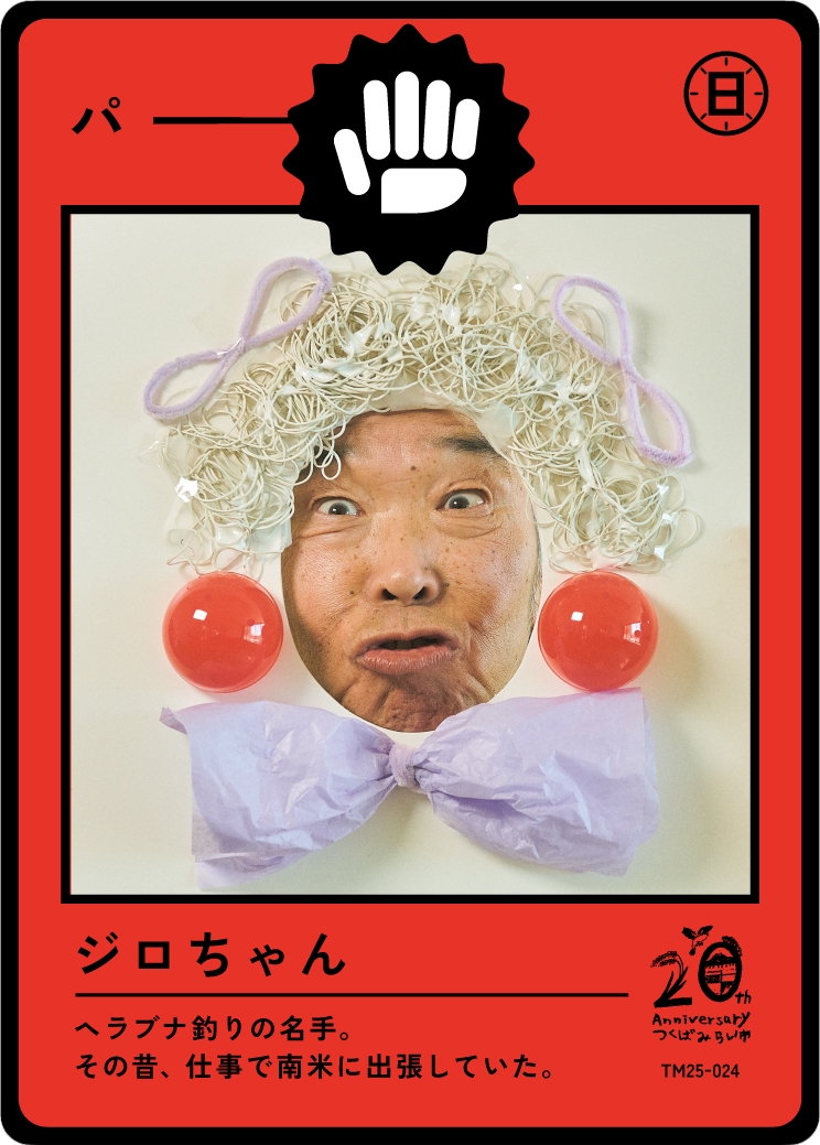 A person with a surprised facial expression is shown through a paper craft clown face with a colorful wig, red cheeks, and a purple bow tie, designed as a playing card with Japanese text and a logo celebrating an anniversary.