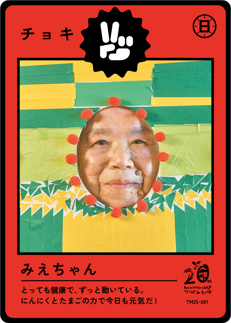 A colorful collage with a person's smiling face at the center, surrounded by red pom-poms, on a background of green and yellow paper. Japanese text and symbols are around the image, indicating a celebration or special card.