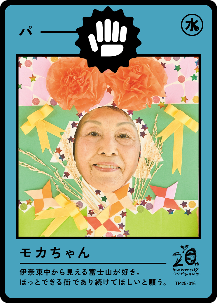 A person’s face is framed within a colorful, handmade paper craft resembling a Japanese trading card, with floral and star decorations, and a black symbol with a white hand at the top. The card has Japanese text and celebrates a 20th anniversary.