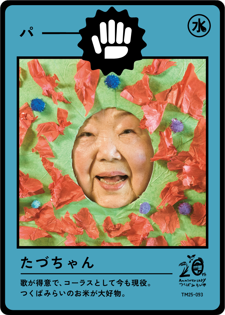 A woman with a big smile poking her head through a colorful craft project decorated with crinkled red paper and small purple and blue pom-poms, resembling a fun, creative face or mask.