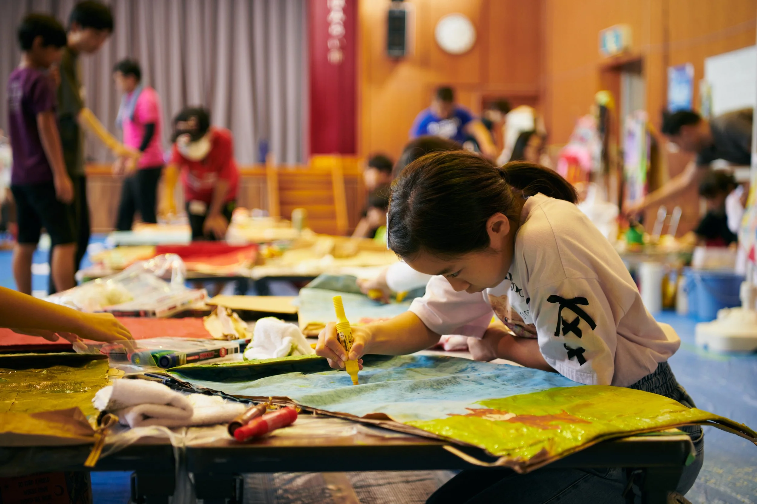 A young girl is drawing or coloring on a large piece of paper on a table surrounded by other children and adults in a room.