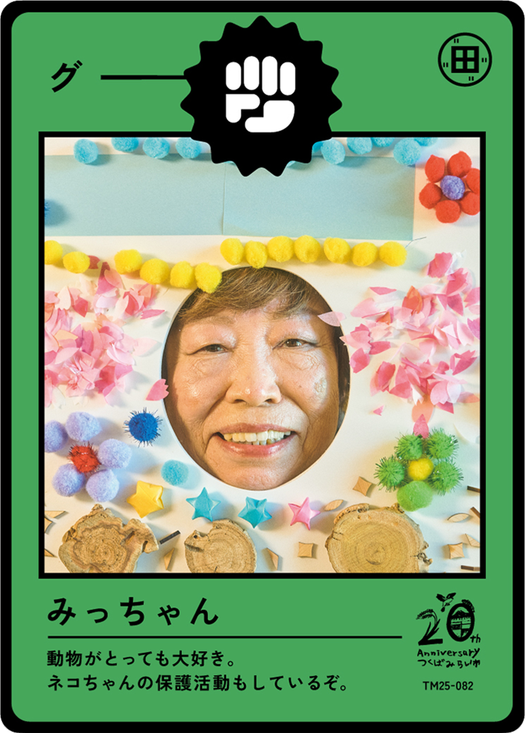 A person with a large, round cutout face in the center of a colorful, decorated frame resembling a card or photo booth picture. The frame has various small, colorful craft decorations including pom-poms, paper flowers, and wooden slices. The person i