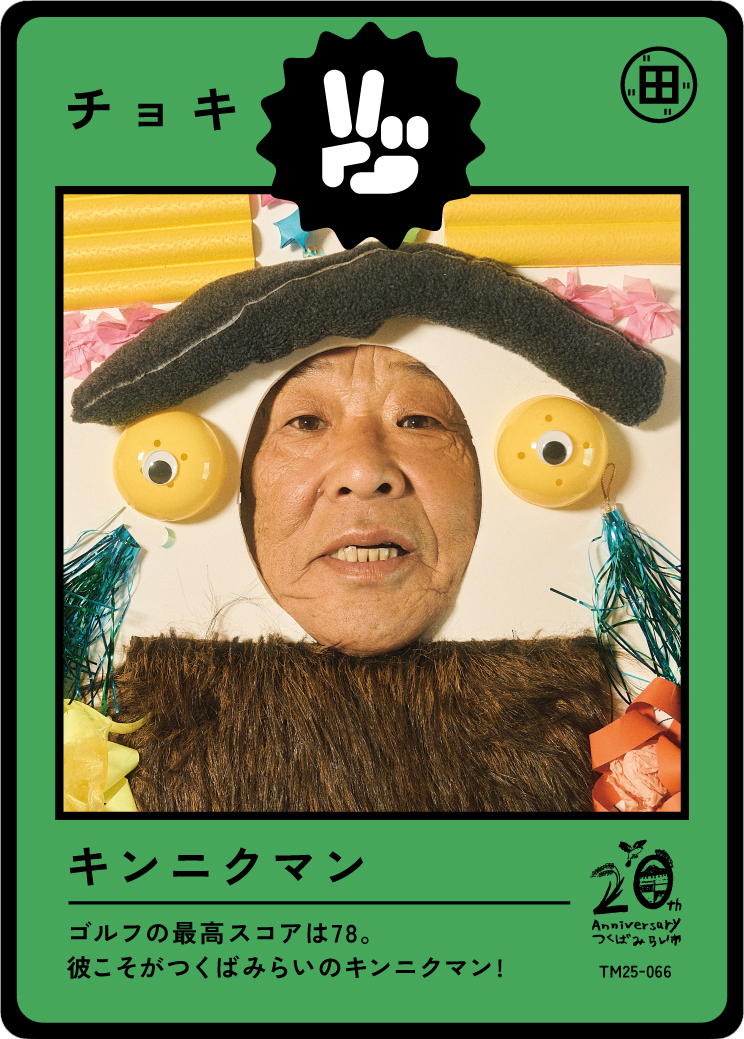 Humanoid figure with a man's face in the center, decorated with a fuzzy hat, yellow objects resembling eyes, and a furry brown fabric around the neck for a costume, framed with a green border that features Japanese text and graphics.
