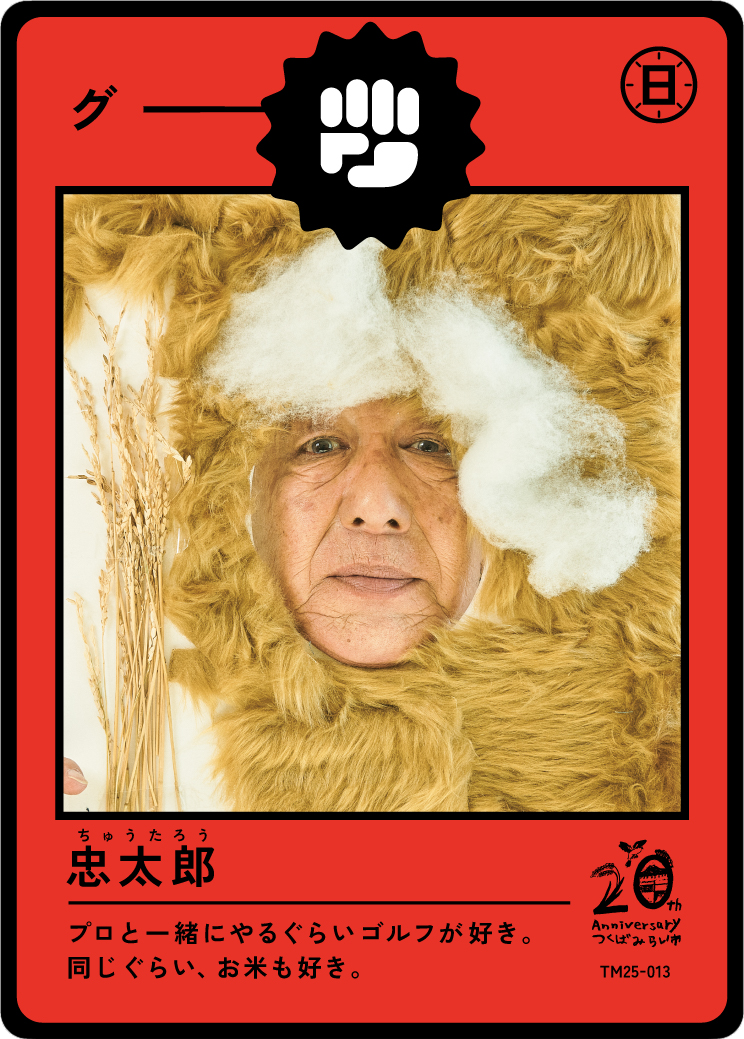 A person with an elderly face is framed by a bear costume with a large bear head and fur. The person is holding a stalk of wheat. The design resembles a trading card with Japanese characters and a 20th anniversary logo.