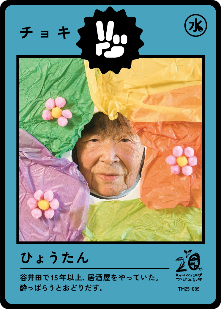 A person with a smiling face surrounded by colorful crumpled tissue paper in green, purple, yellow, and orange, with small pink pom-pom flowers on the tissue paper.