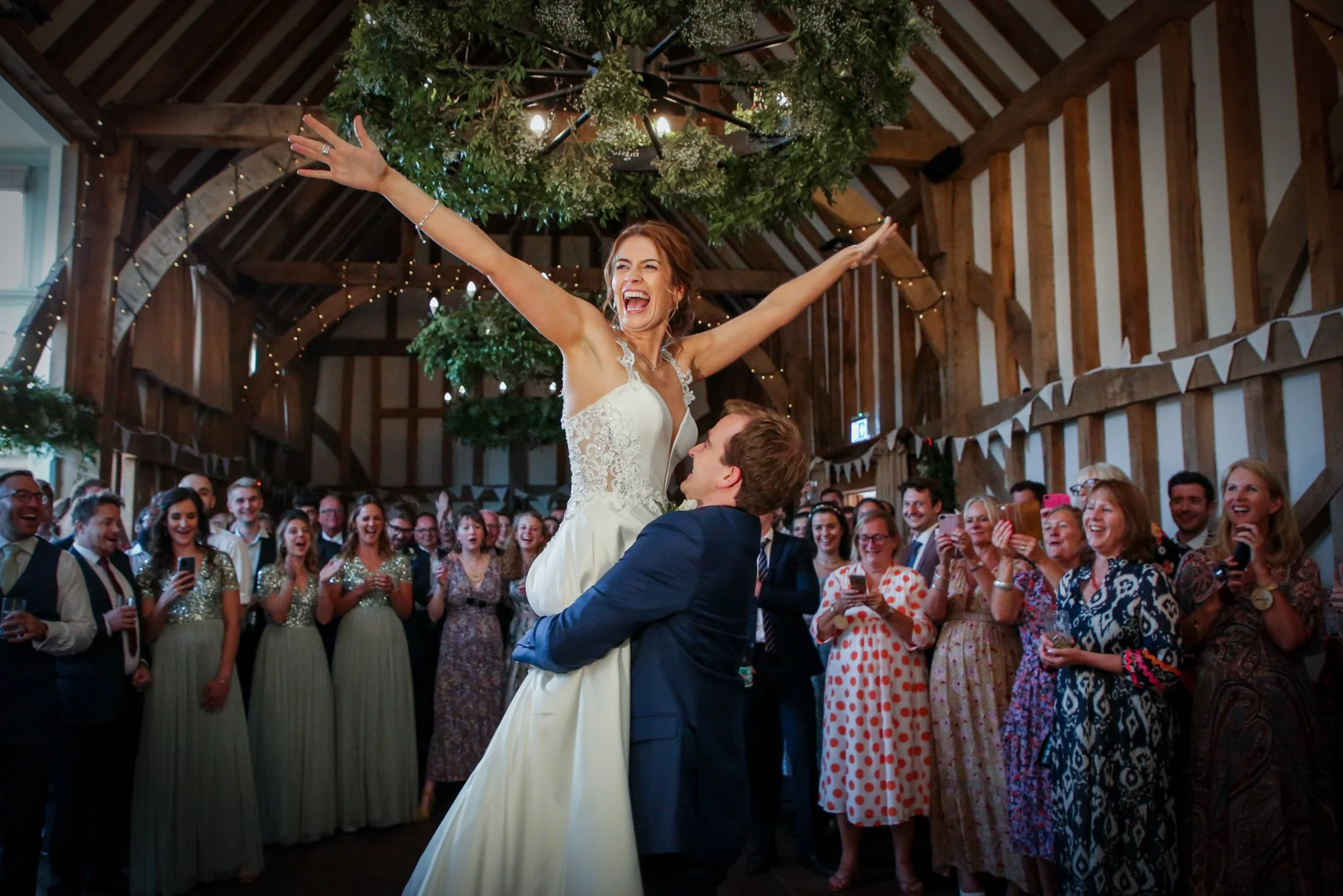Bride and groom dancing at their wedding reception inside a wooden barn, surrounded by smiling guests, with festive lights and greenery on the ceiling.