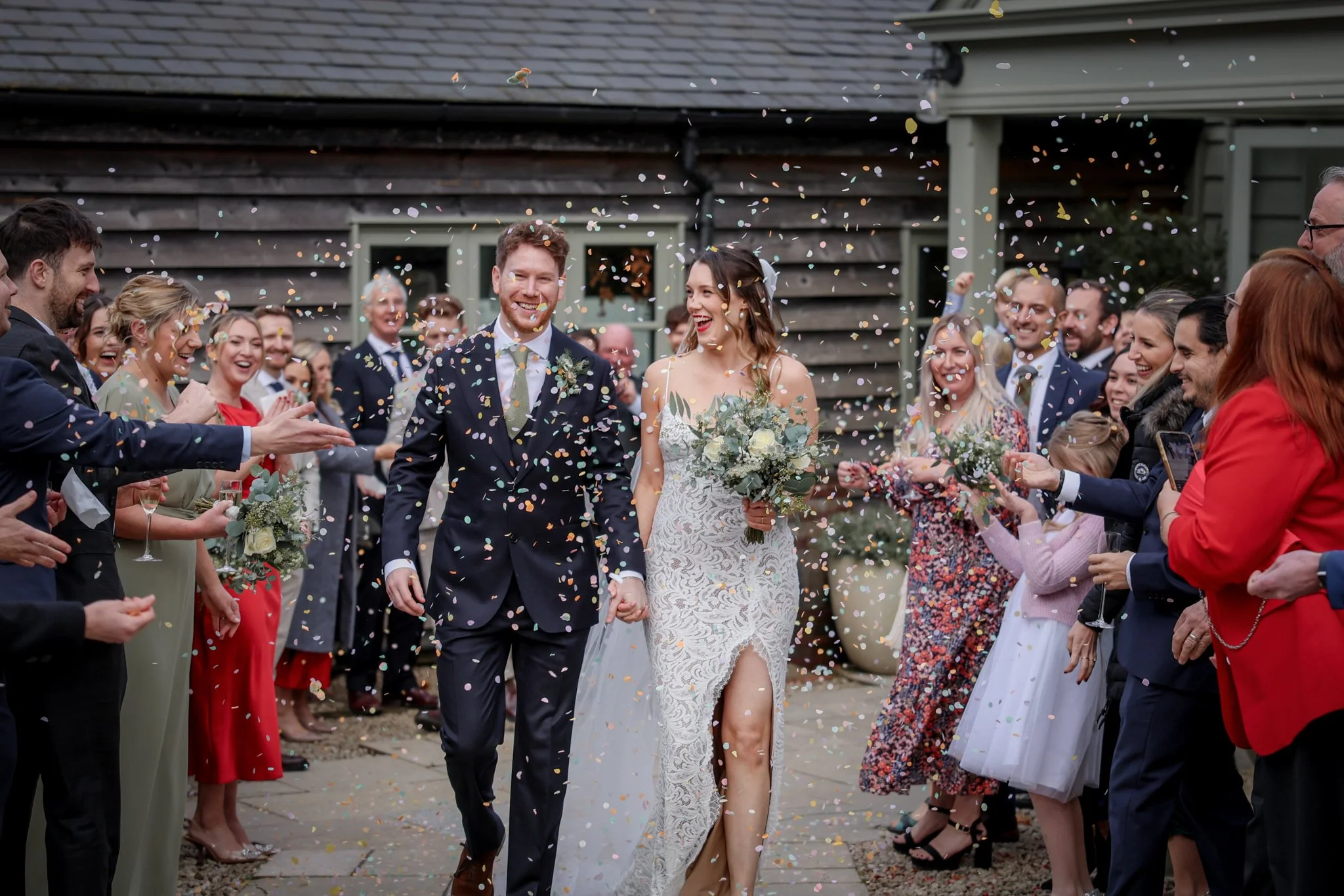 A newlywed couple walks hand in hand through a celebration crowd, surrounded by guests throwing colorful confetti during an outdoor wedding ceremony.