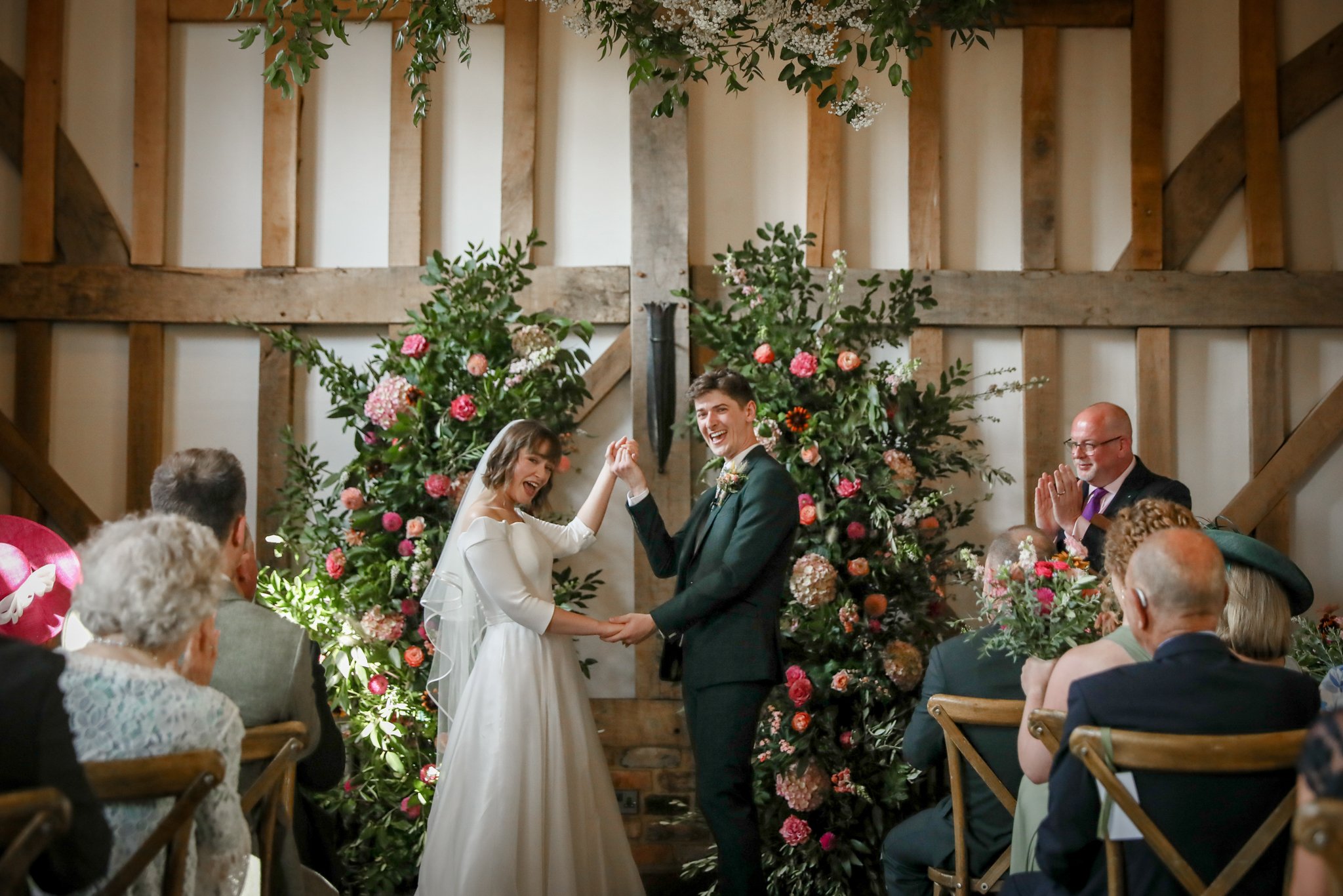 A couple getting married, holding hands and smiling during their wedding ceremony inside a rustic venue with wooden beams and floral decorations.